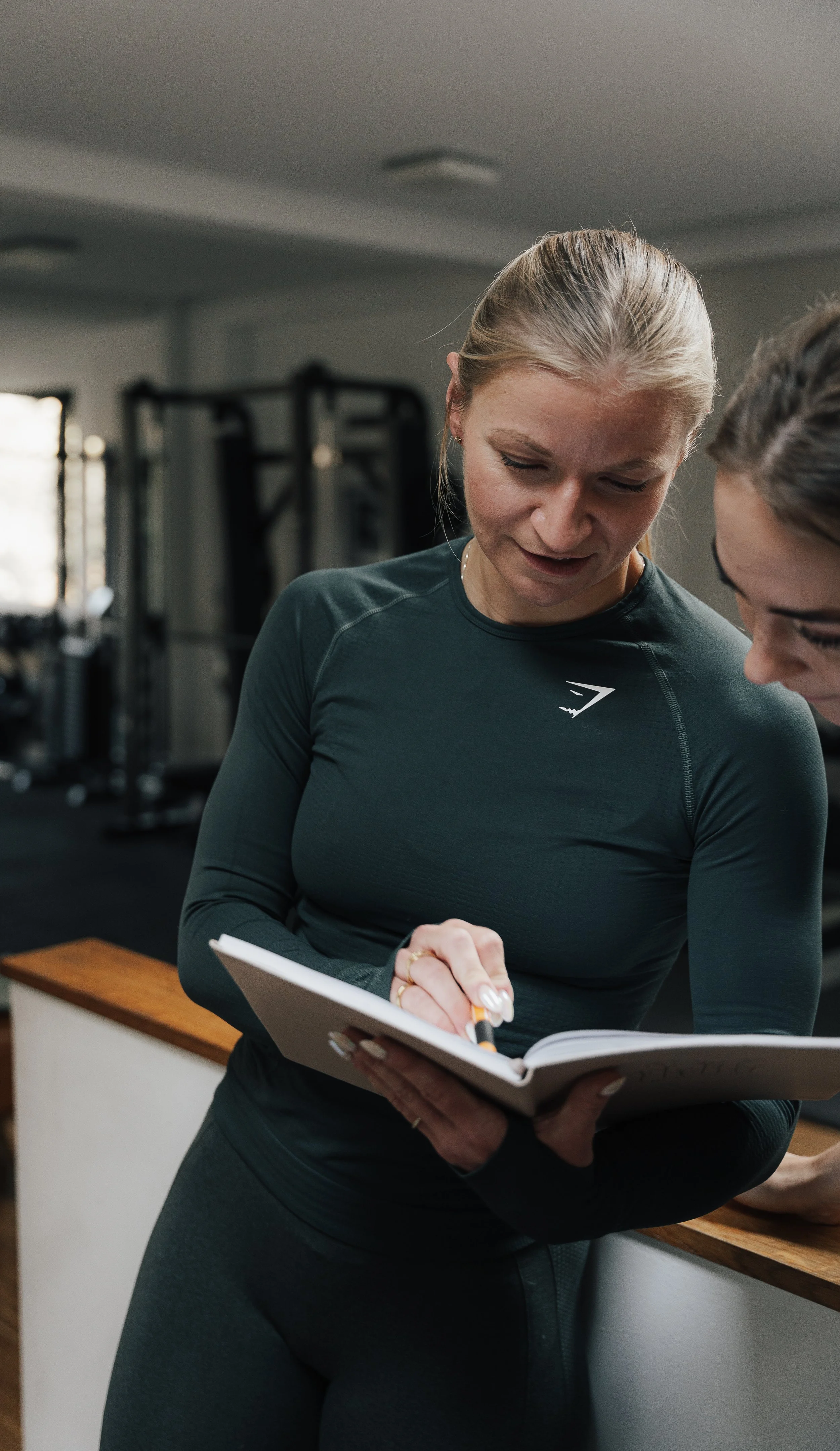 Zwei Frauen im Fitnessstudio, eine schreibt in ein Notizbuch, die andere schaut zu.