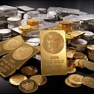 Stacks of silver and gold coins with a gold bar in the center labeled '9999 Fine Gold 10 Troy Ounces'.