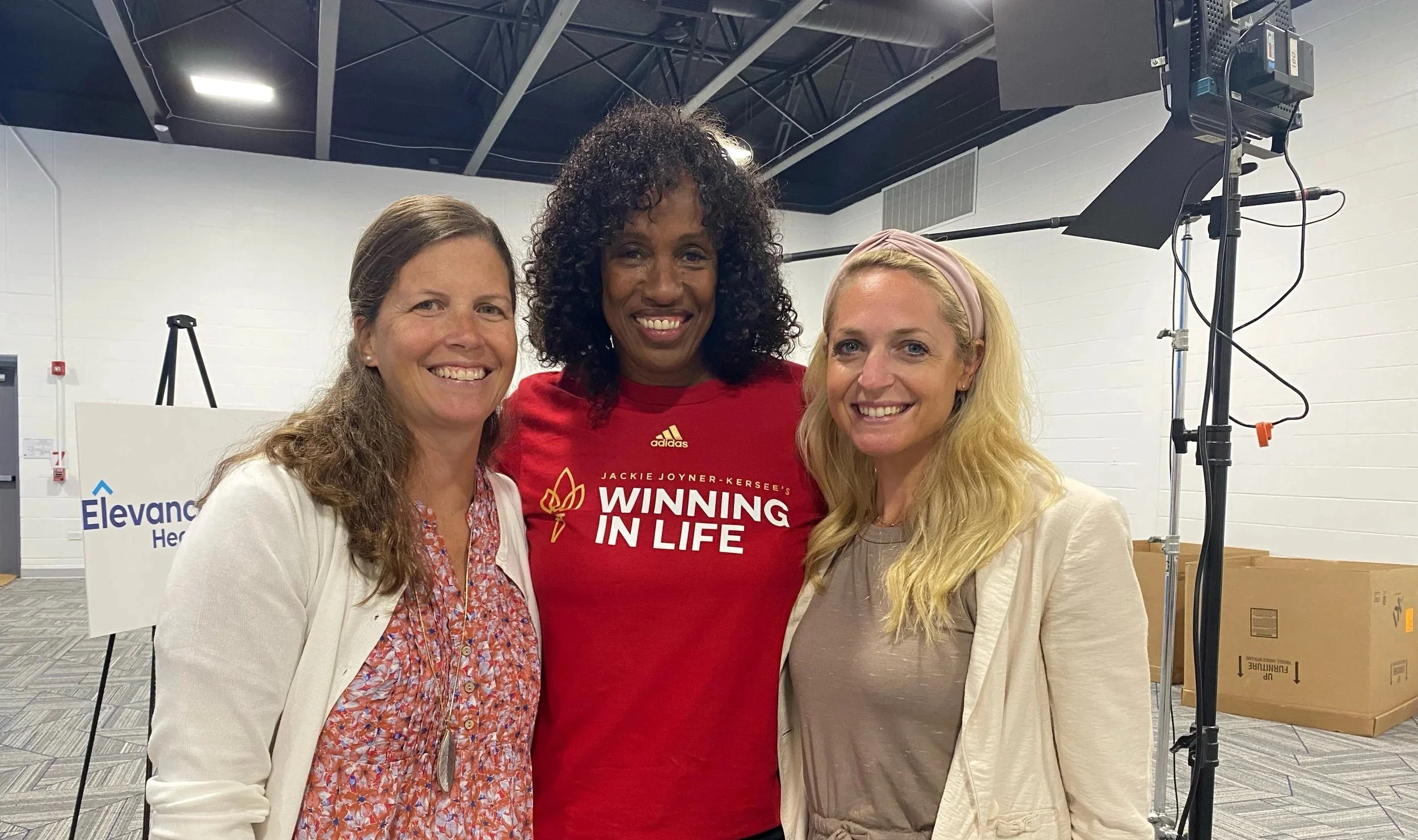 Three women smiling and standing close together in an indoor setting, with two women wearing white blazers and one woman in a red T-shirt that reads 'Winning in Life'.