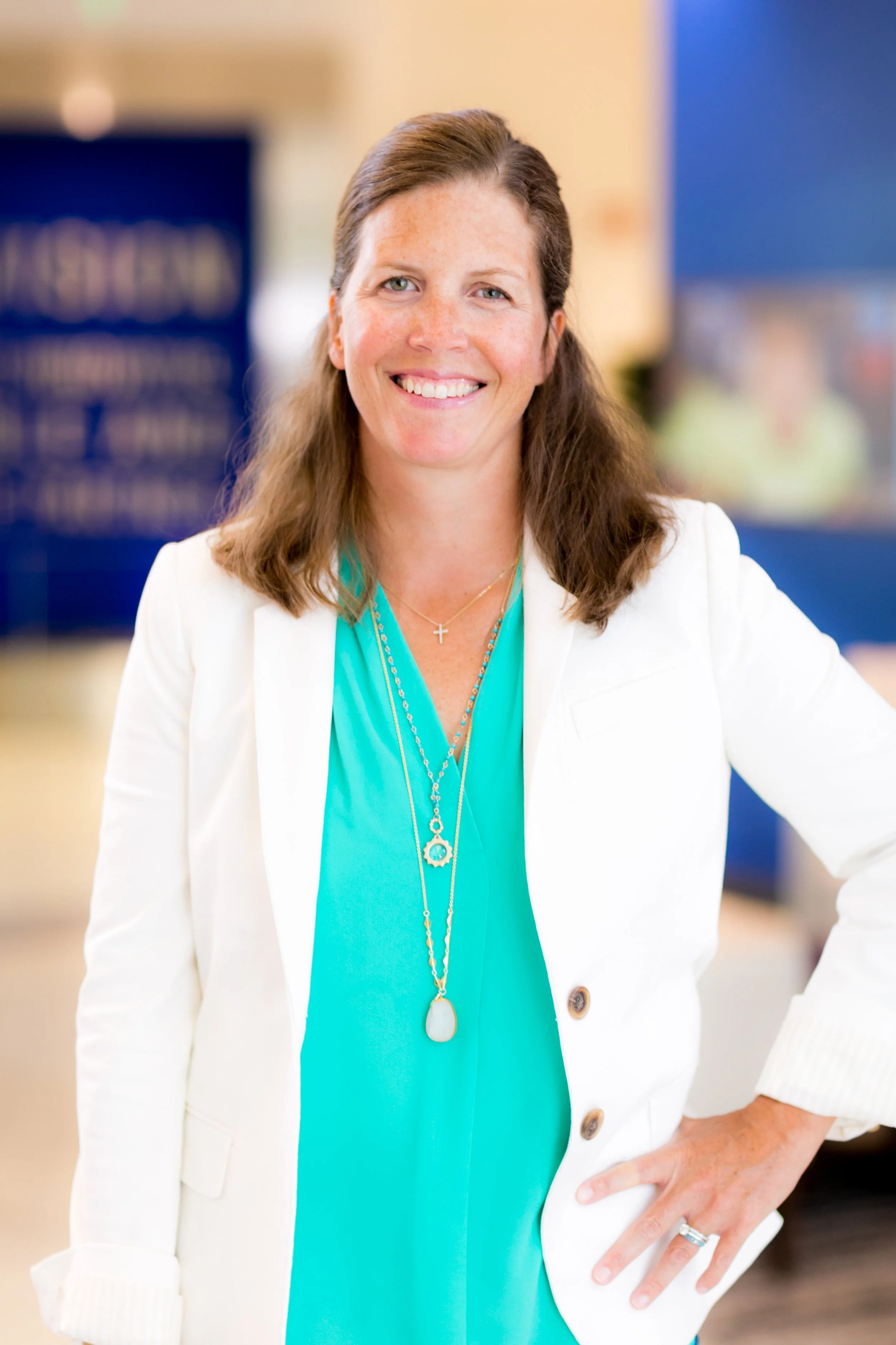 A woman smiling, wearing a white blazer over a turquoise top, standing indoors.
