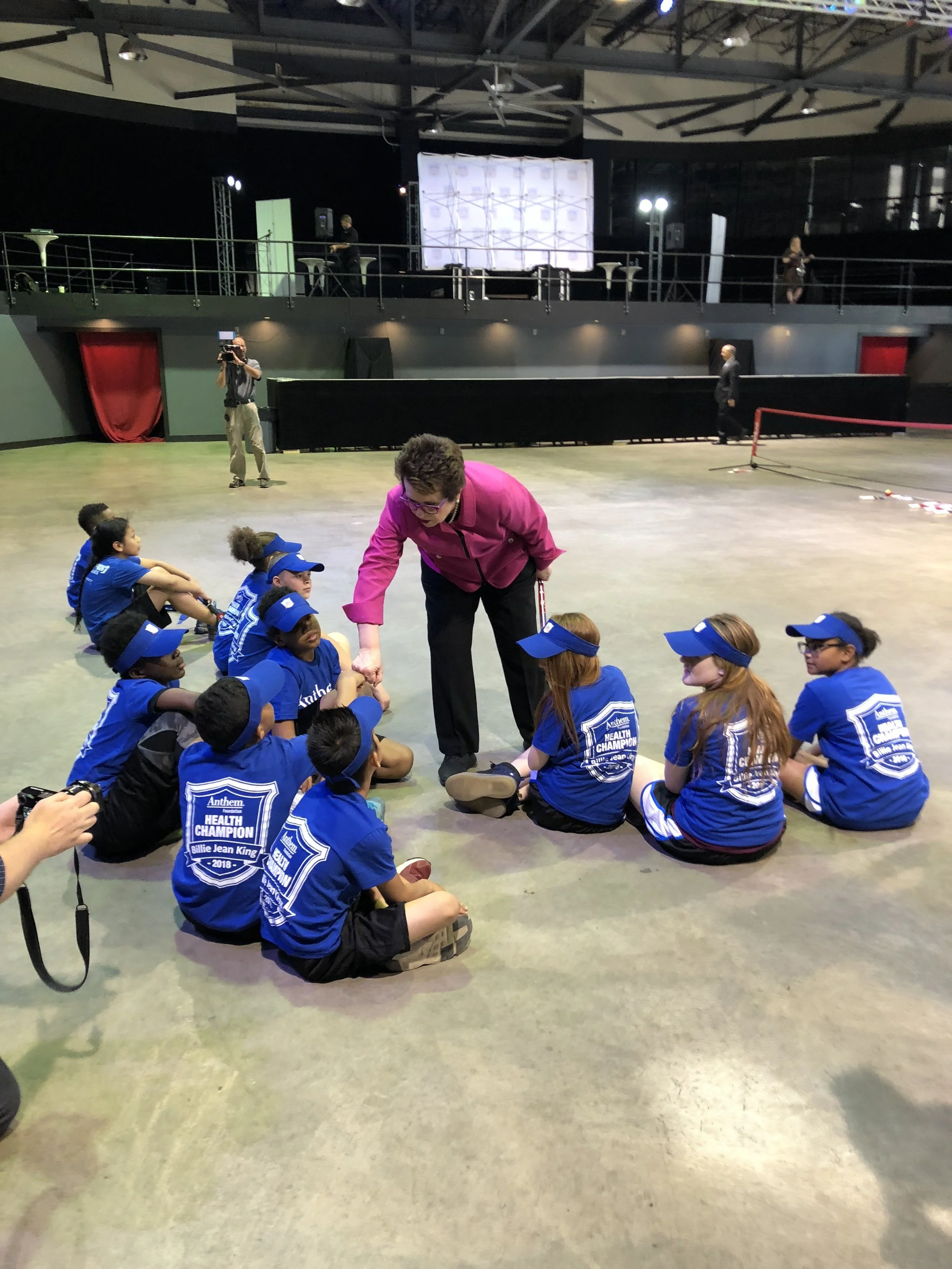 A woman in a pink jacket is sitting on the floor and talking to children wearing blue shirts and caps, sitting in a semi-circle indoors.