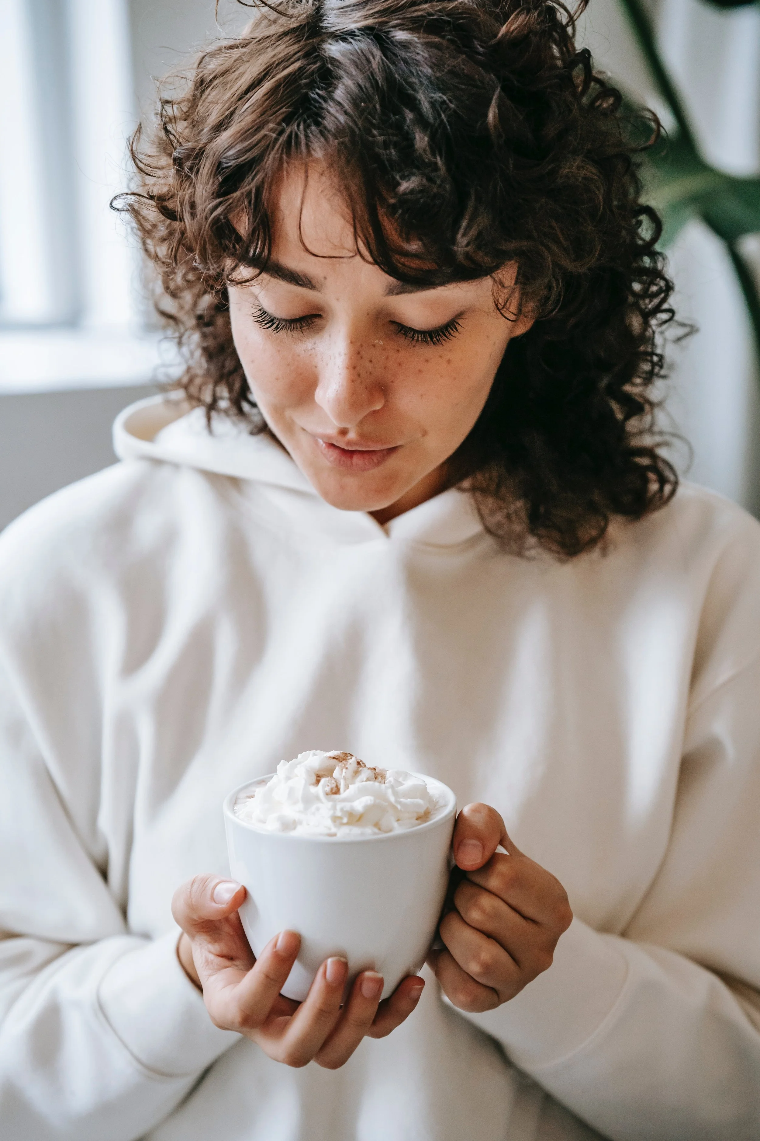 A young woman with curly brown hair and freckles, wearing a white hoodie, holds a white mug topped with whipped cream and sprinkled with cinnamon or nutmeg, looking down at the drink with a gentle smile.