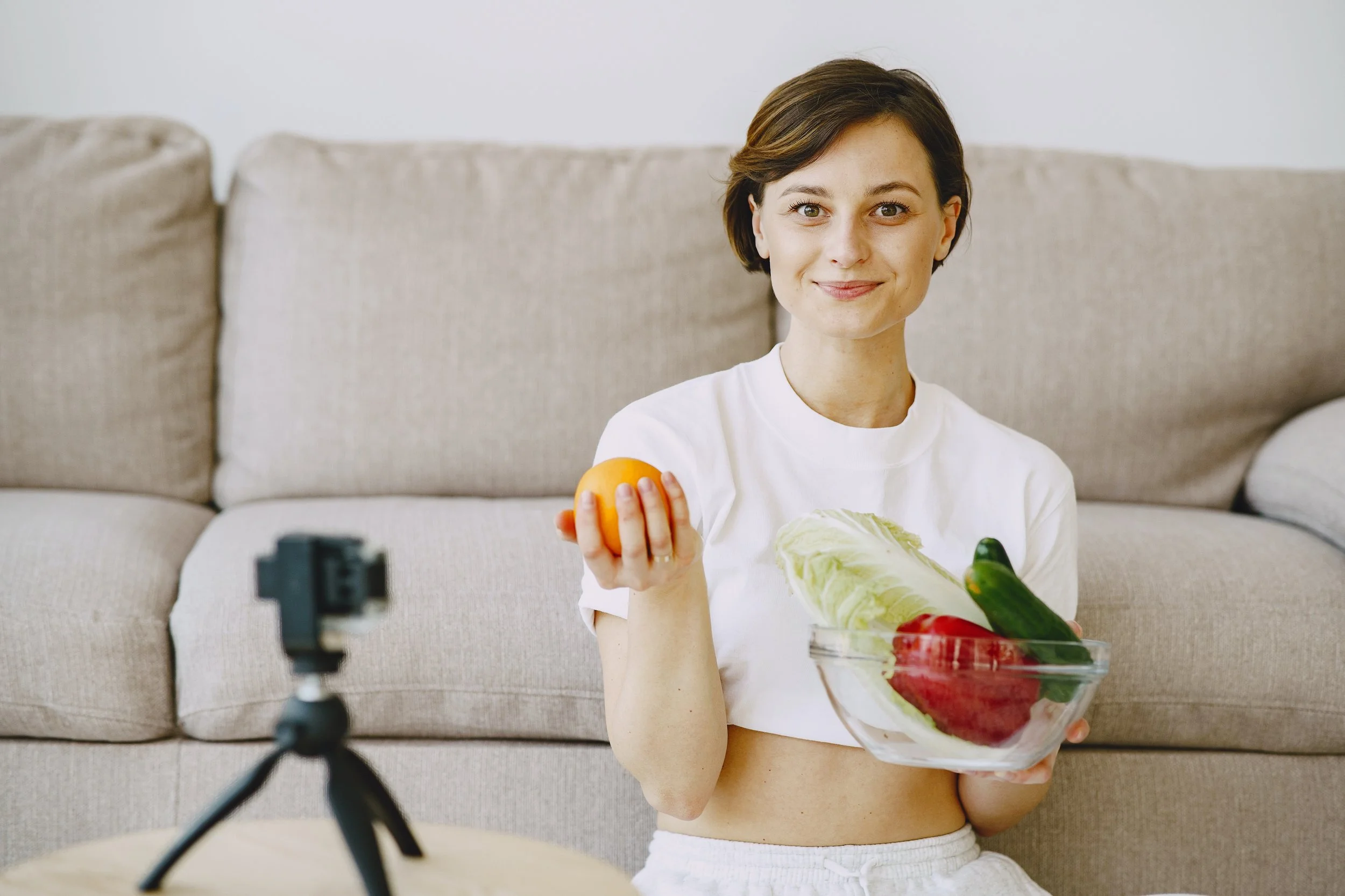 A woman with short brown hair and a white shirt sits on a beige couch holding a glass bowl of vegetables and an orange, while being recorded by a camera on a tripod.