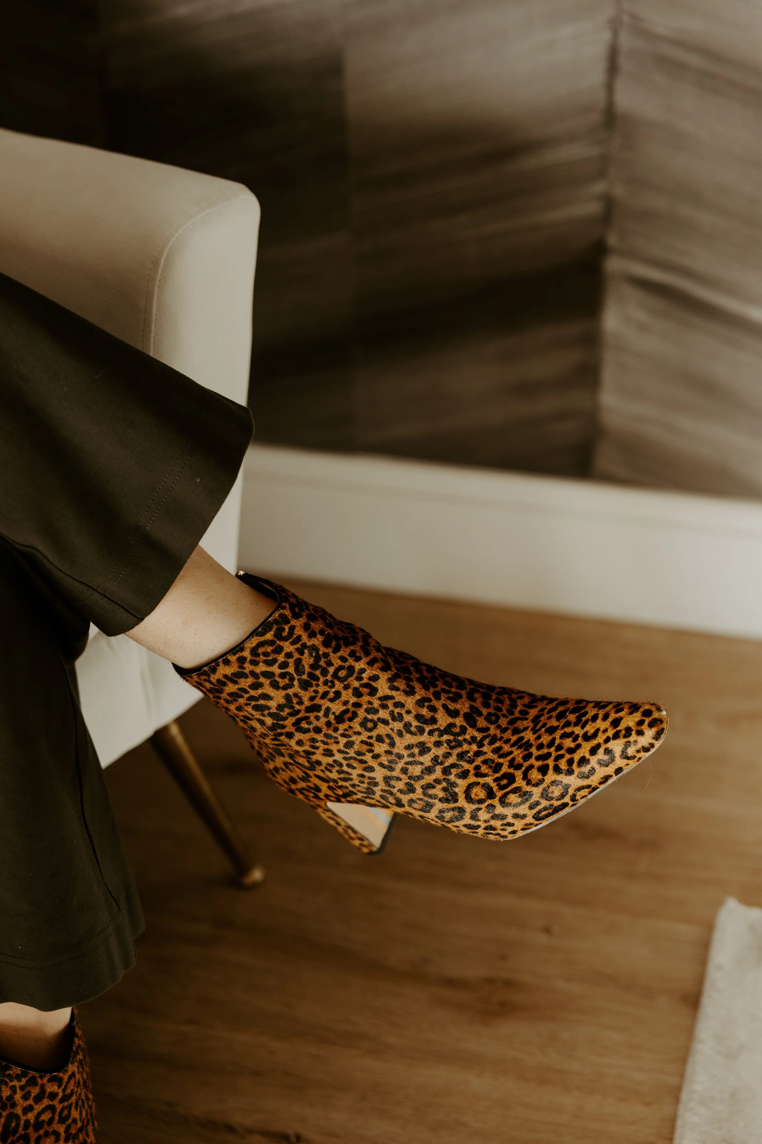 A person sitting on a metal chair with only their legs visible, wearing light blue jeans, white socks, and white sneakers, holding a closed book against a plain white wall.