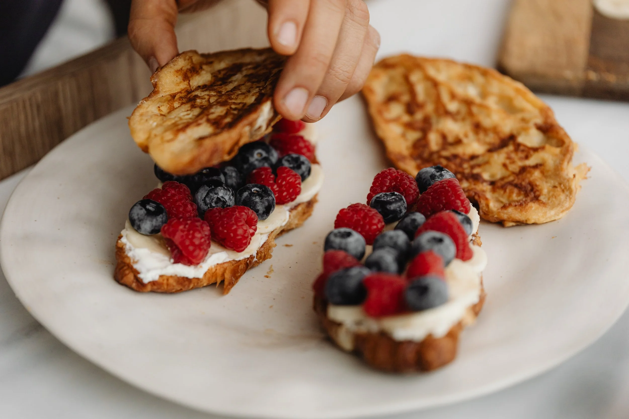 Person placing toasted bread with cream cheese and fresh berries on a white plate.