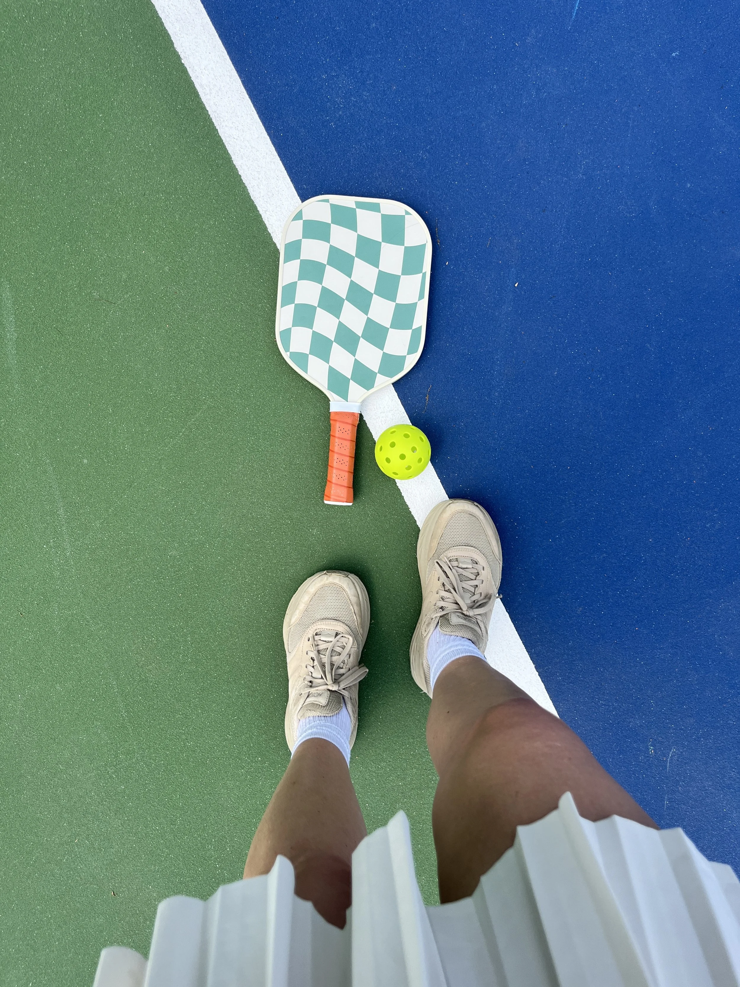 Tennis court with a paddle, ball, and a person's legs in sneakers and white socks.