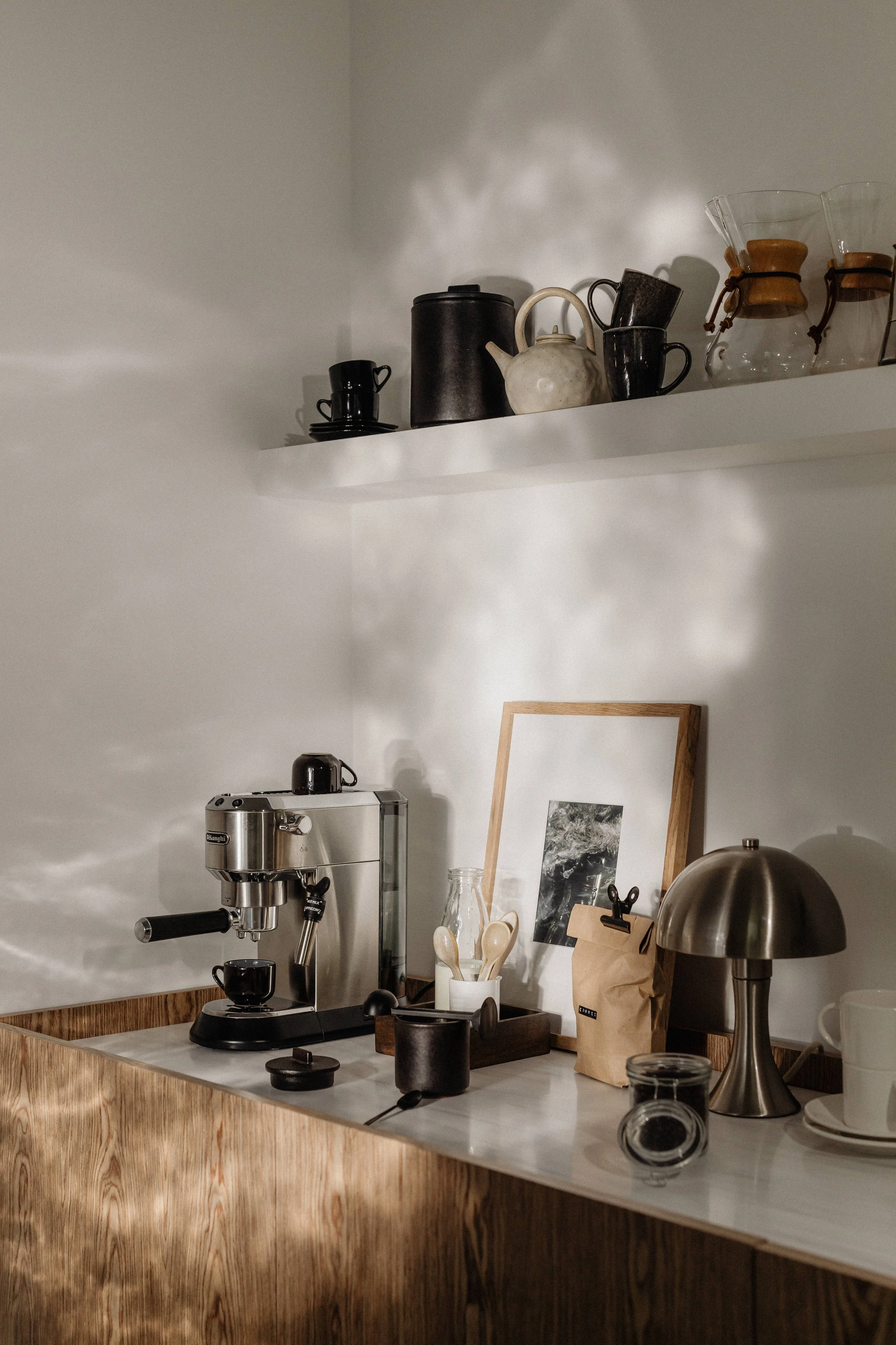 Coffee station with a modern espresso machine, framed photo, mug, jar, and lamp on a wooden countertop; shelves with teapots, cups, and coffee makers on the white wall.