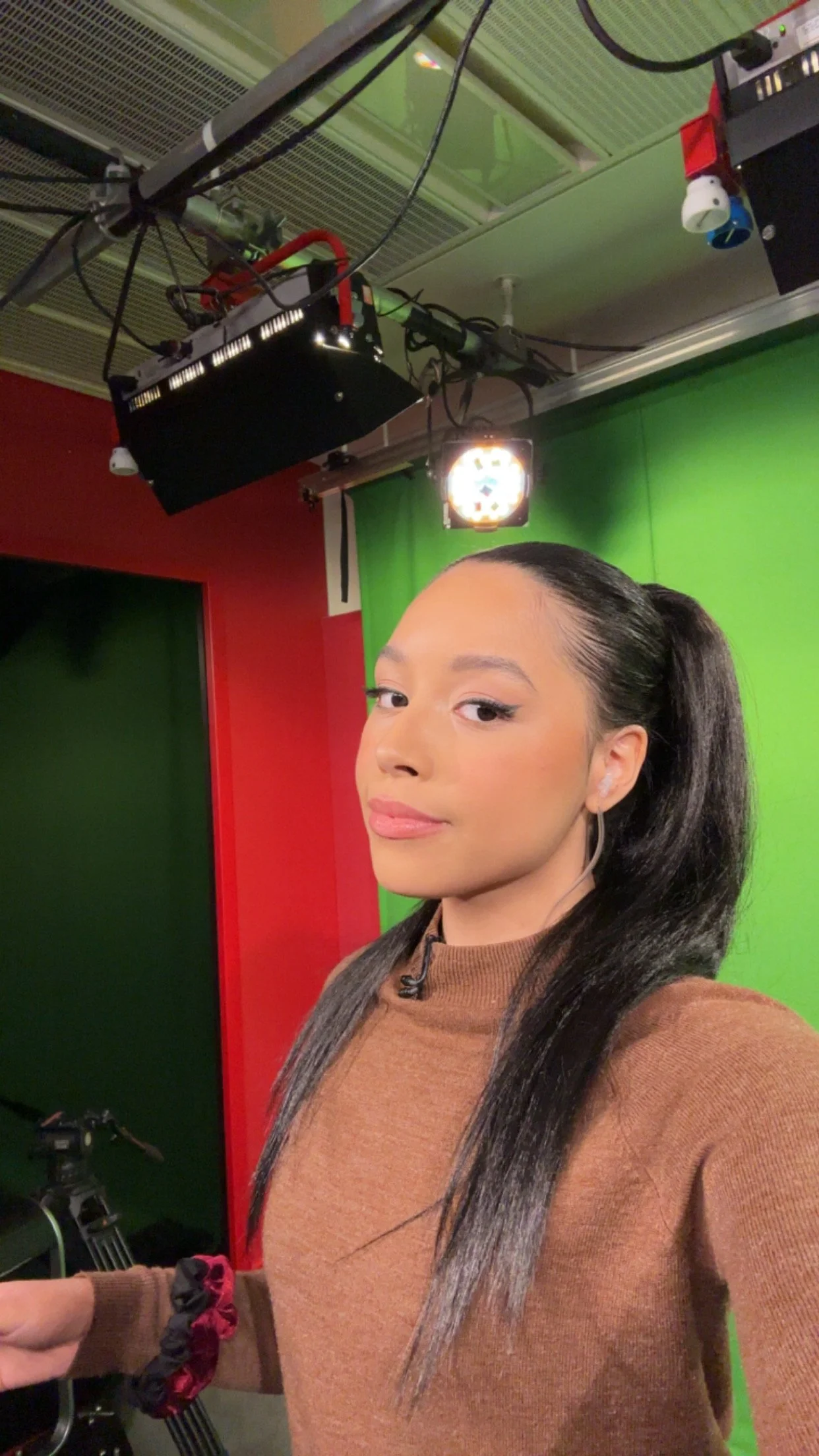 A woman in a brown sweater taking a selfie in a studio with a green screen background, camera equipment overhead, and a video camera on a tripod in the background.