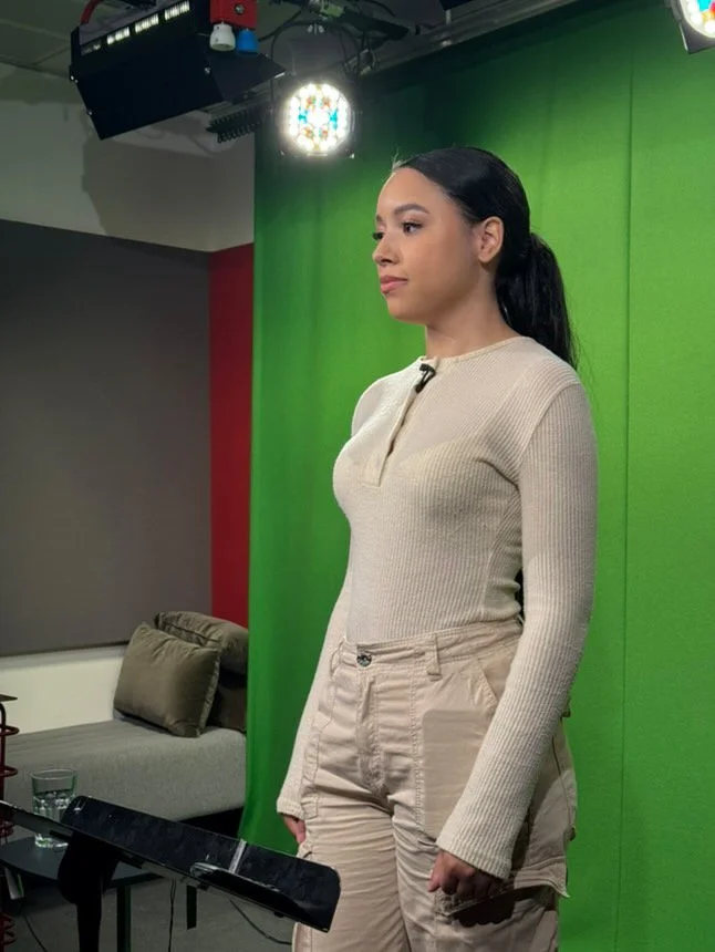 A woman with dark hair tied back standing in front of a green screen in a studio, with studio lights overhead and a sofa in the background.