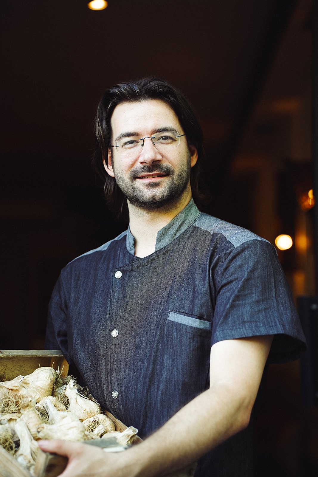 Homme souriant portant une veste de chef et tenant des têtes d'ail, intérieur avec éclairage chaud.