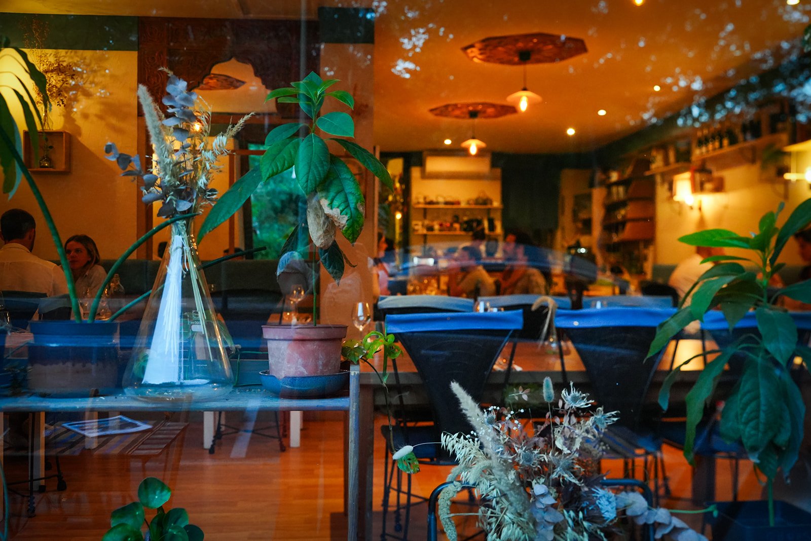 Une vue de l'intérieur d'un restaurant ou d'un café, vue à travers une fenêtre avec plusieurs plantes en pot sur une étagère devant la vitre, et des clients assis à des tables à l'intérieur.