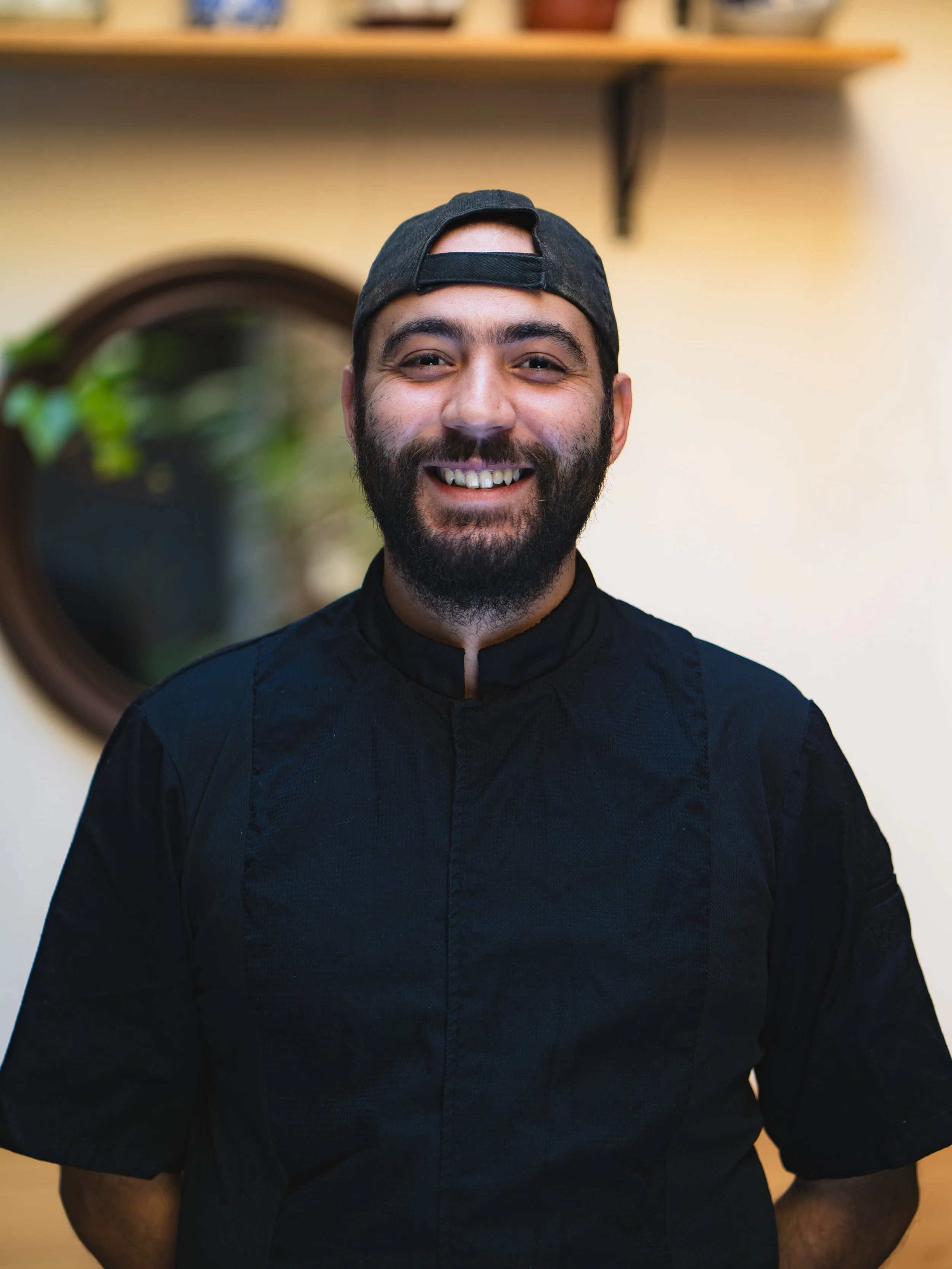 Un homme souriant portant une veste de chef et une casquette noire, dans une cuisine