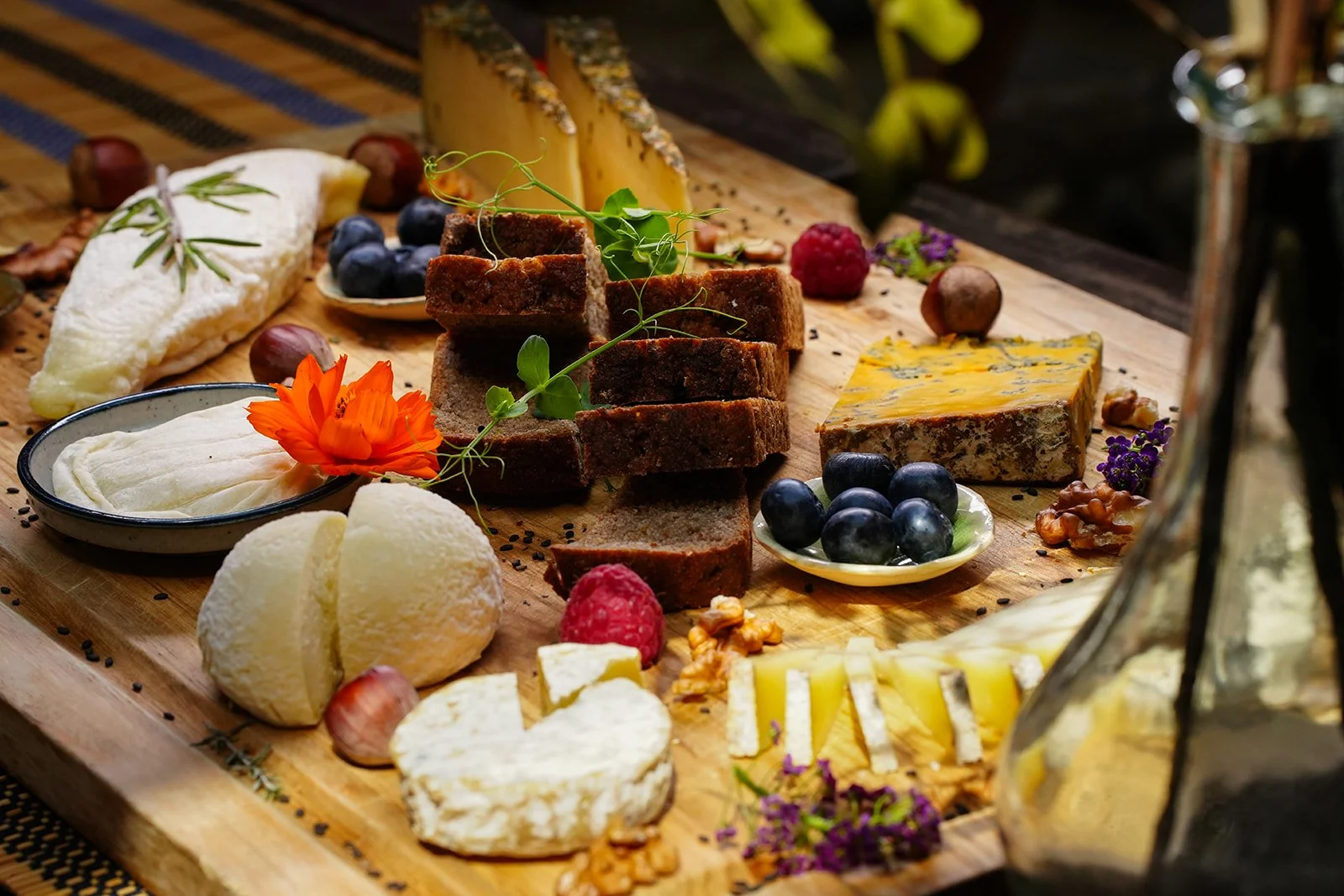 Plateau de fromages variés, fruits, noix, et pain sur une planche en bois, décoré avec des fleurs et des garnitures colorées.