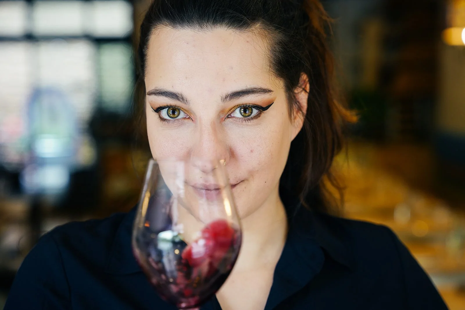 Une femme regarde au travers d'un verre de vin rouge avec un fond flou d'un intérieur de restaurant.