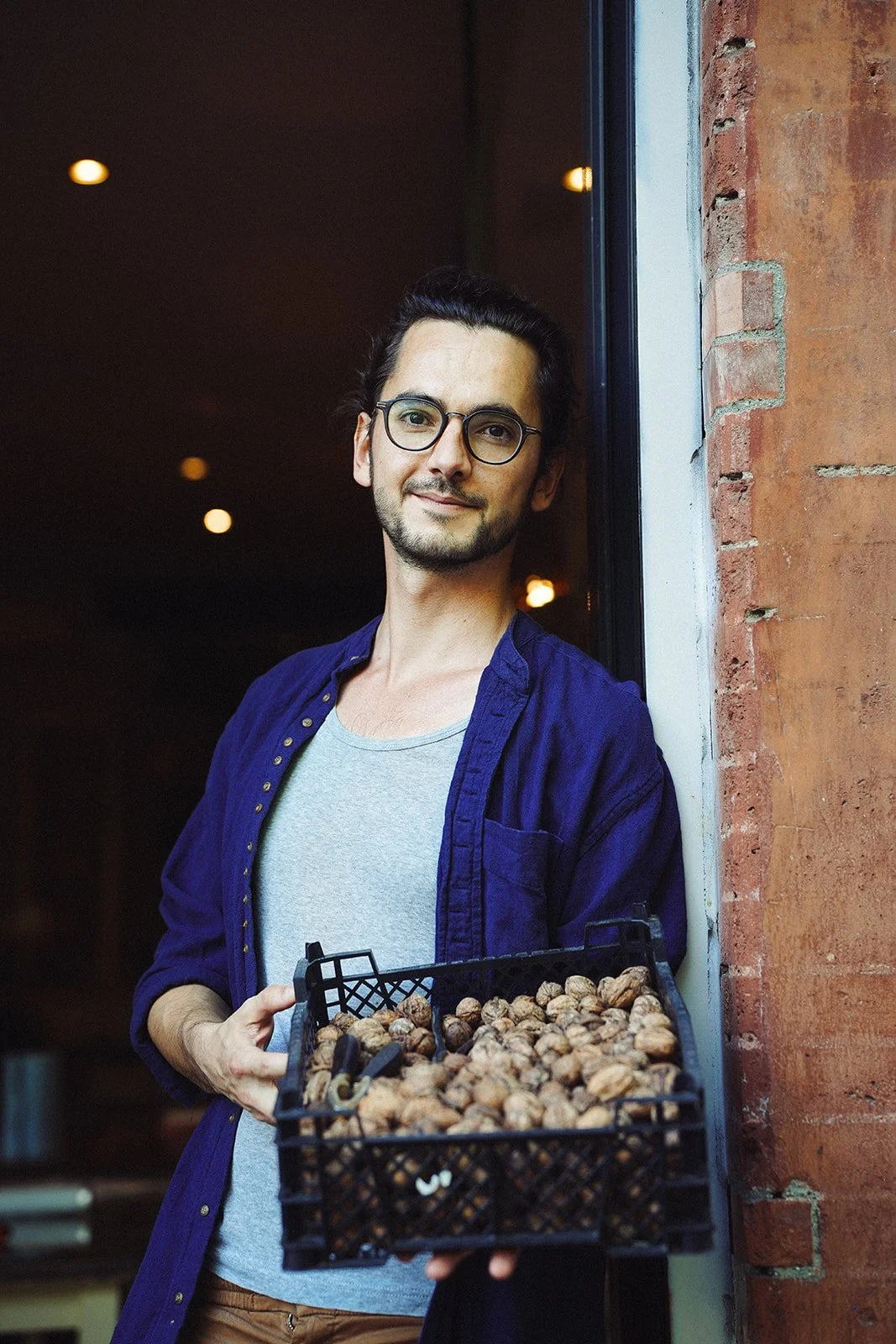 Jeune homme avec lunettes tenant un plateau de noix, debout à côté d'une fenêtre dans un bâtiment en briques.