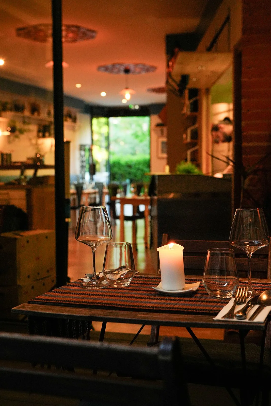 Table de restaurant avec une bougie allumée, deux verres à vin, un verre à eau, une fourchette et une cuillère, dans un restaurant intérieur avec lumière tamisée et vue sur un extérieur verdoyant.