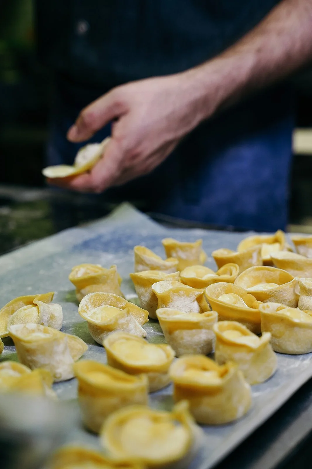 Gros plan sur des tortellinis crus disposés sur une plaque recouverte de papier sulfurisé, avec une main en arrière préparant ou façonnant des pâtes.