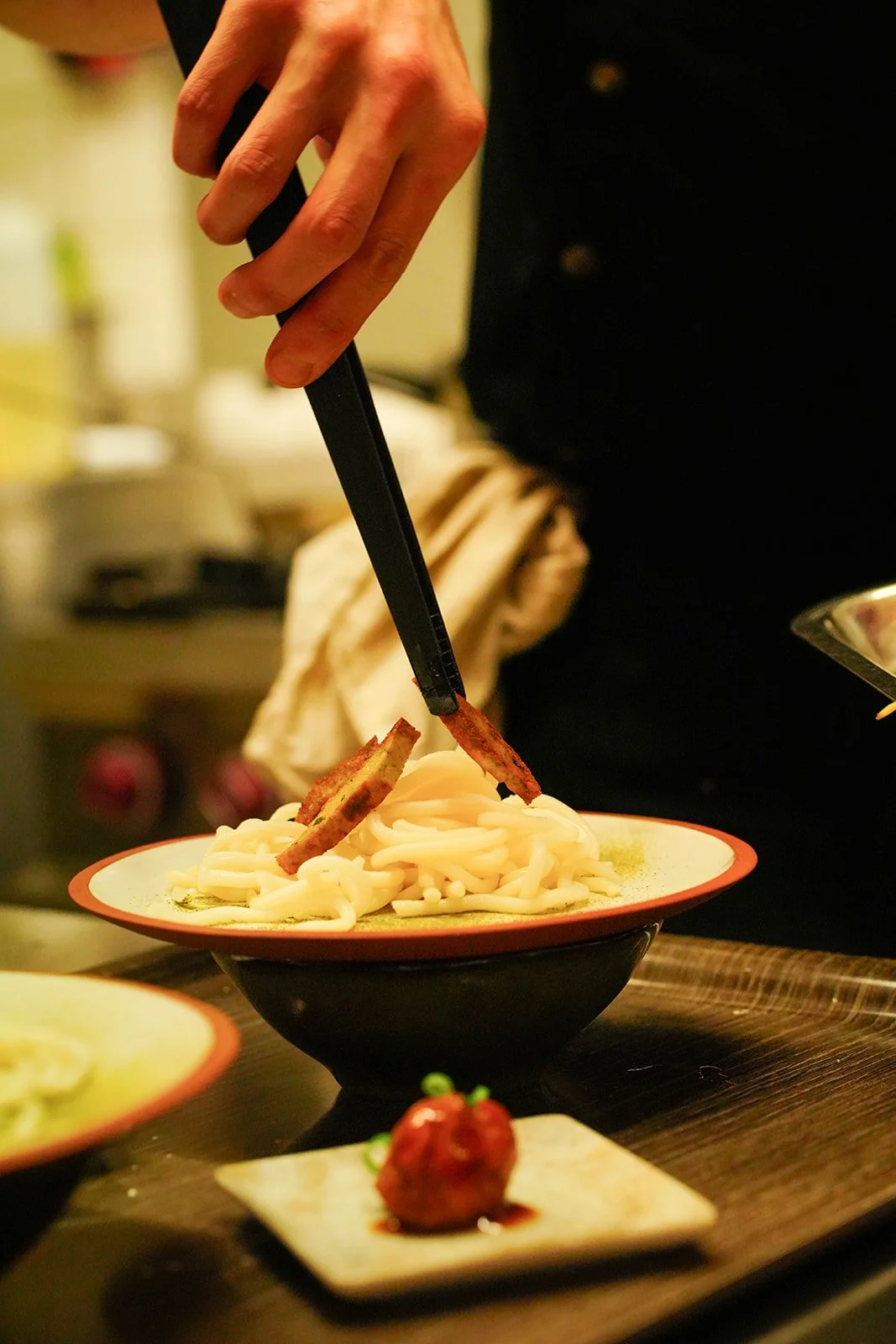 Person utilisant des pinces pour déposer des tranches de viande sur un plat de nouilles, avec une petite assiette de sauce à côté.