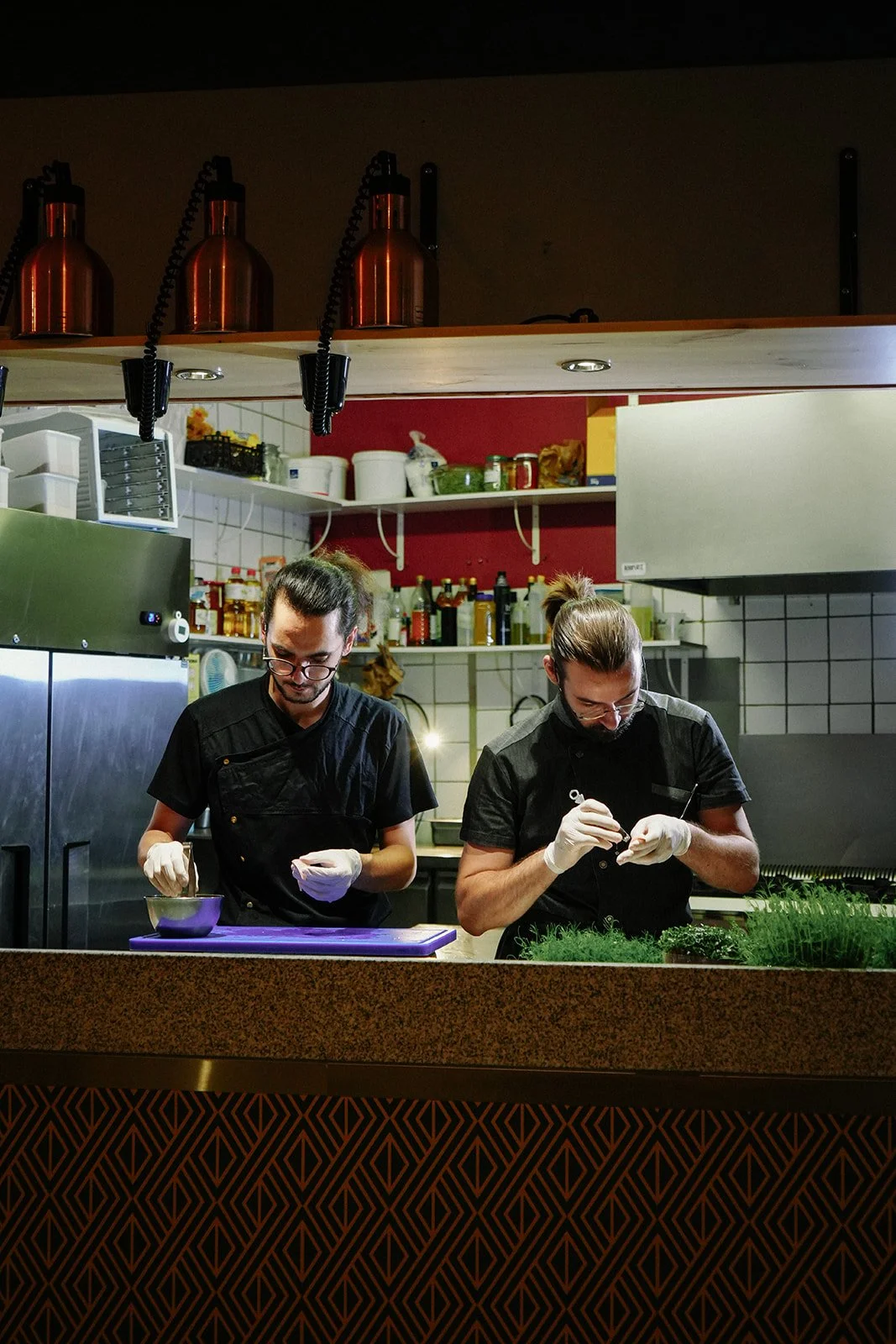 Deux chefs en train de préparer des plats dans une cuisine de restaurant, portant des gants en latex blanc, avec des étagères remplies d'épices et d'ustensiles en arrière-plan.
