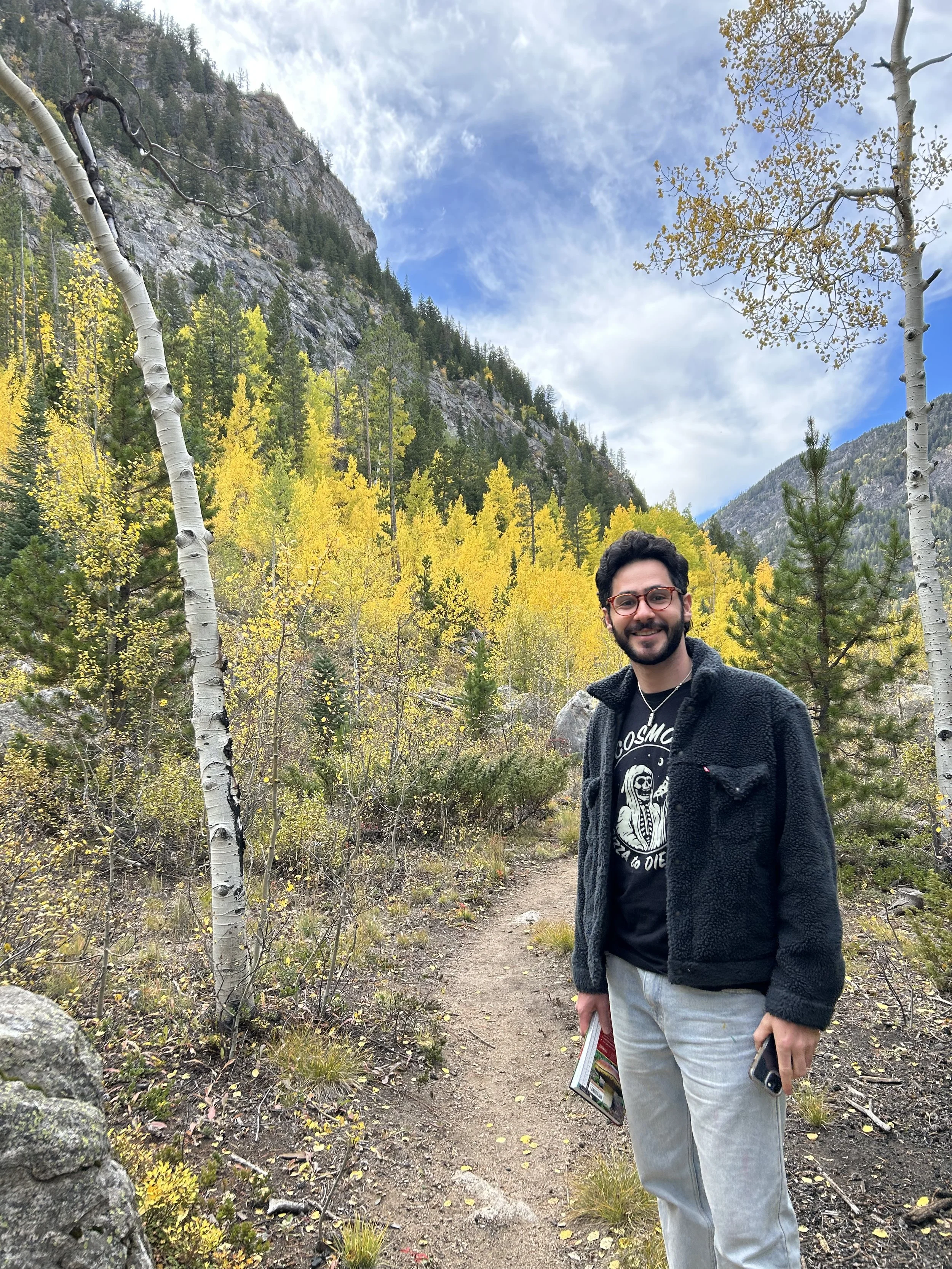 A man with glasses and a beard smiling while standing on a trail in a mountainous forest with yellow and green foliage and a blue sky with clouds.