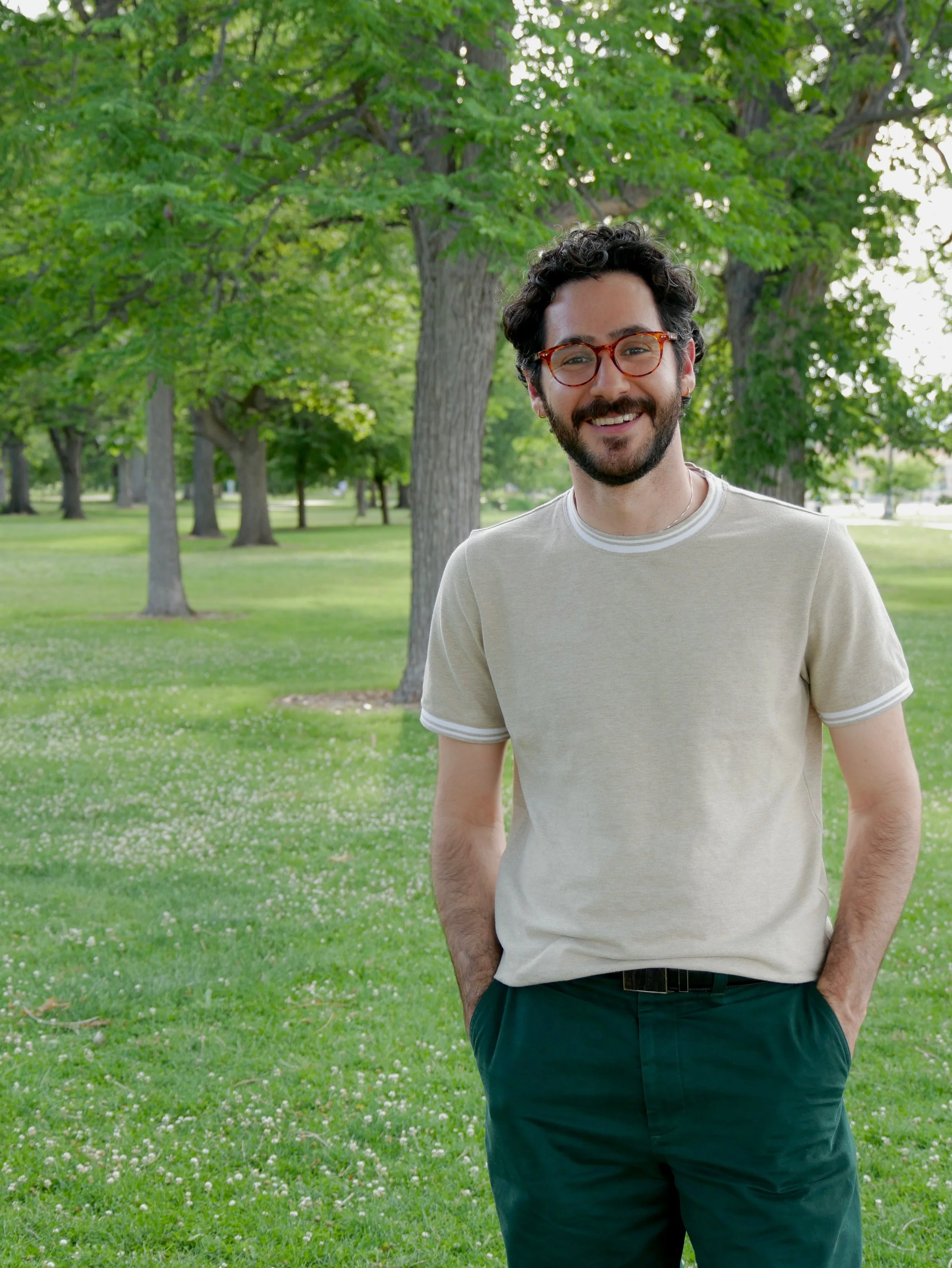 A man with curly dark hair, glasses with orange frames, and a beard is smiling while standing in a park with green trees and grass.