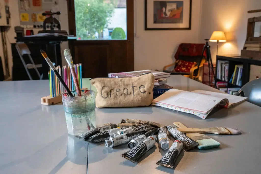 An art and study area with tubes of black and white paint, paintbrushes in a glass jar, colored pencils, an open book, and a sign that says 'Create' on a cloth pouch, with a living room in the background.