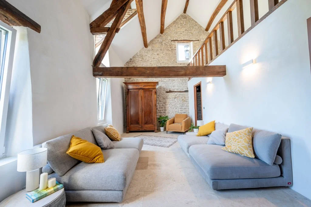 Living room with high vaulted ceiling, exposed beams, stone wall, and white walls. Two light gray sofas with yellow and gray pillows, a small side table with a lamp, a beige armchair, and a wooden wardrobe. There are large windows allowing natural li