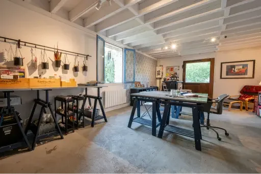 An art studio with a large table, drawing materials, a workbench with hanging art supplies, and artwork on the walls.