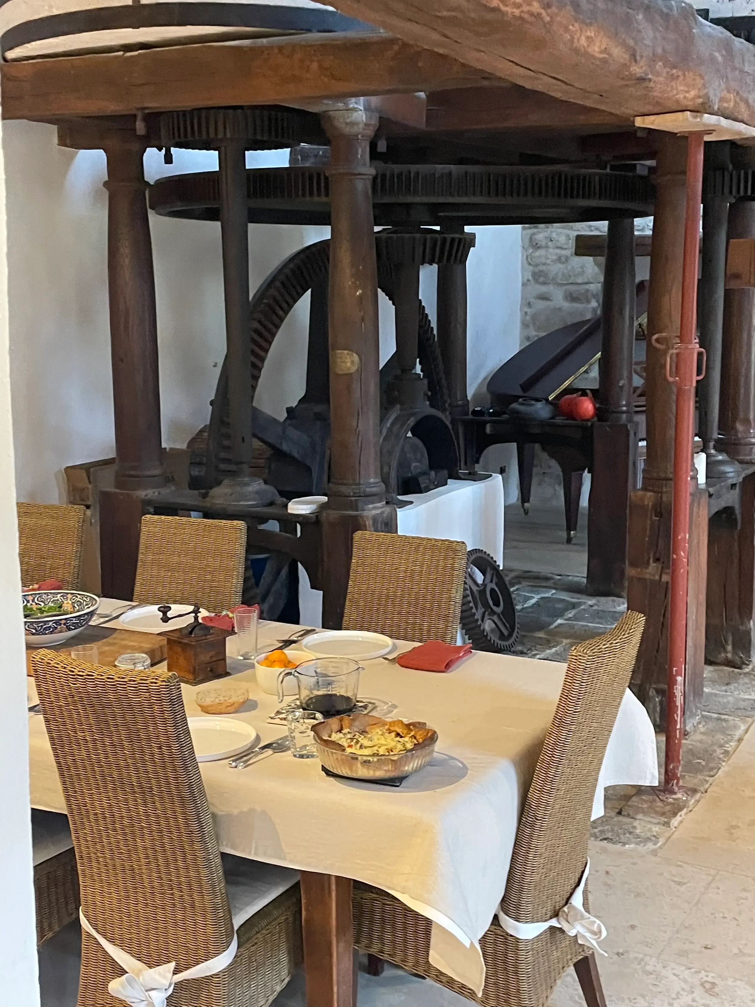 A dining room with a table set for a meal, featuring a casserole dish, wine glass, and plates. Old machinery and large gears are visible in the background, with a wooden and stone decor.