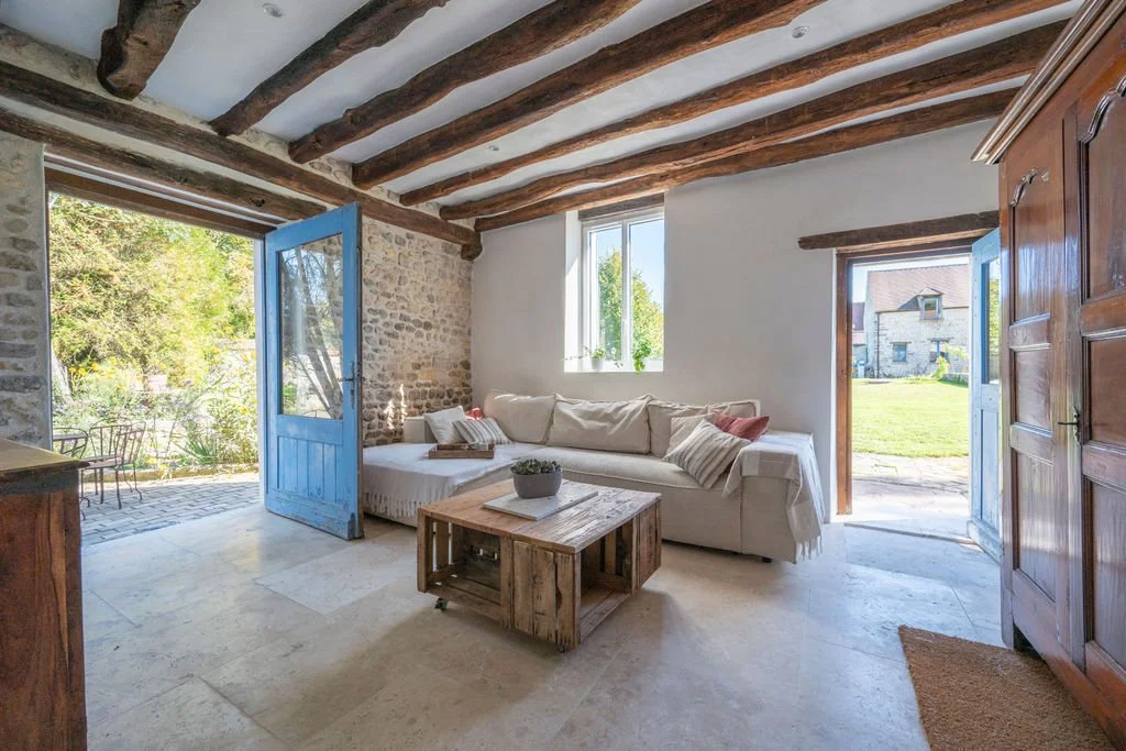 Living room with white walls, exposed wooden ceiling beams, a white sofa, a rustic wooden coffee table, two open doors leading outside, and a window allowing natural light, with a stone exterior visible outside.