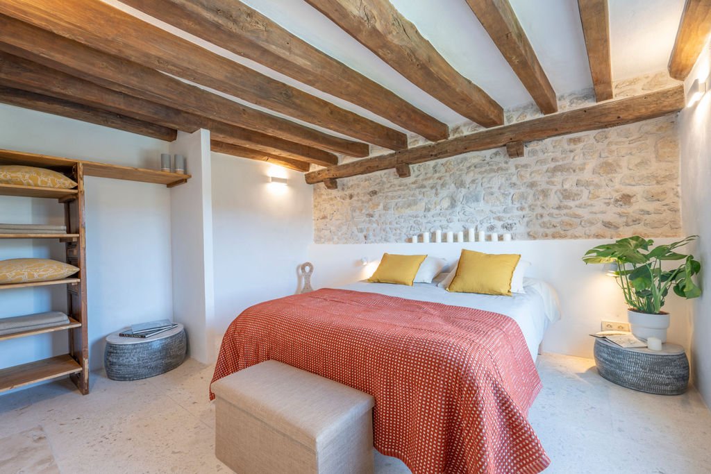 A cozy bedroom with a bed featuring a white headboard, yellow pillows, and a red polka dot blanket. The room has a stone wall behind the bed, wooden ceiling beams, a potted plant on a bedside table, and a beige ottoman at the foot of the bed.