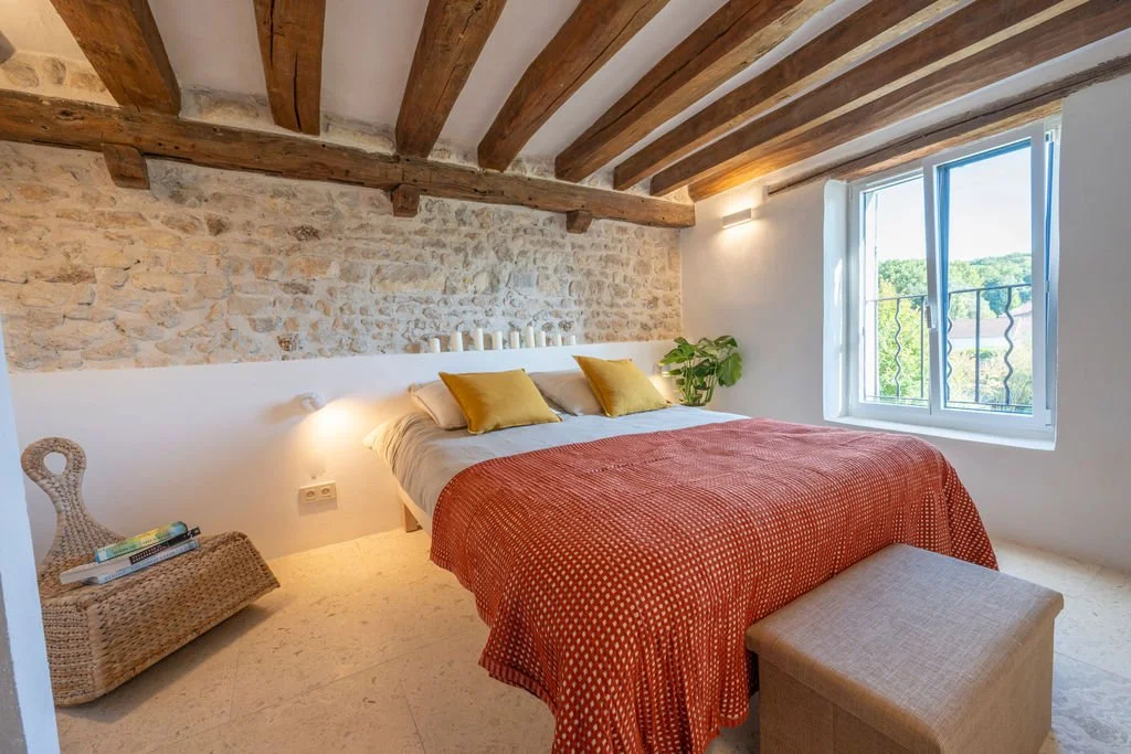 A cozy bedroom with a large bed, neutral bedding, two yellow pillows, a woven bench at foot, natural light from a window, exposed wooden beams on the ceiling, a stone accent wall, a potted plant, and a bedside lamp.