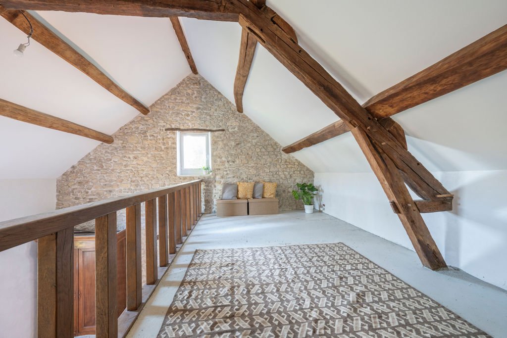 An attic space with exposed wooden beams, a small window, a beige couch with pillows, a potted plant, and a patterned area rug on a light-colored floor.