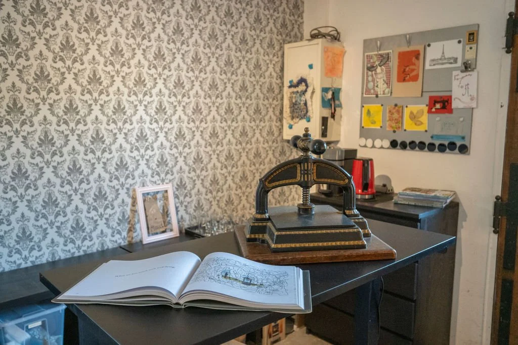 A vintage hand-crank press on a black table with an open coloring book, a small picture frame, and various decorative items in the background.