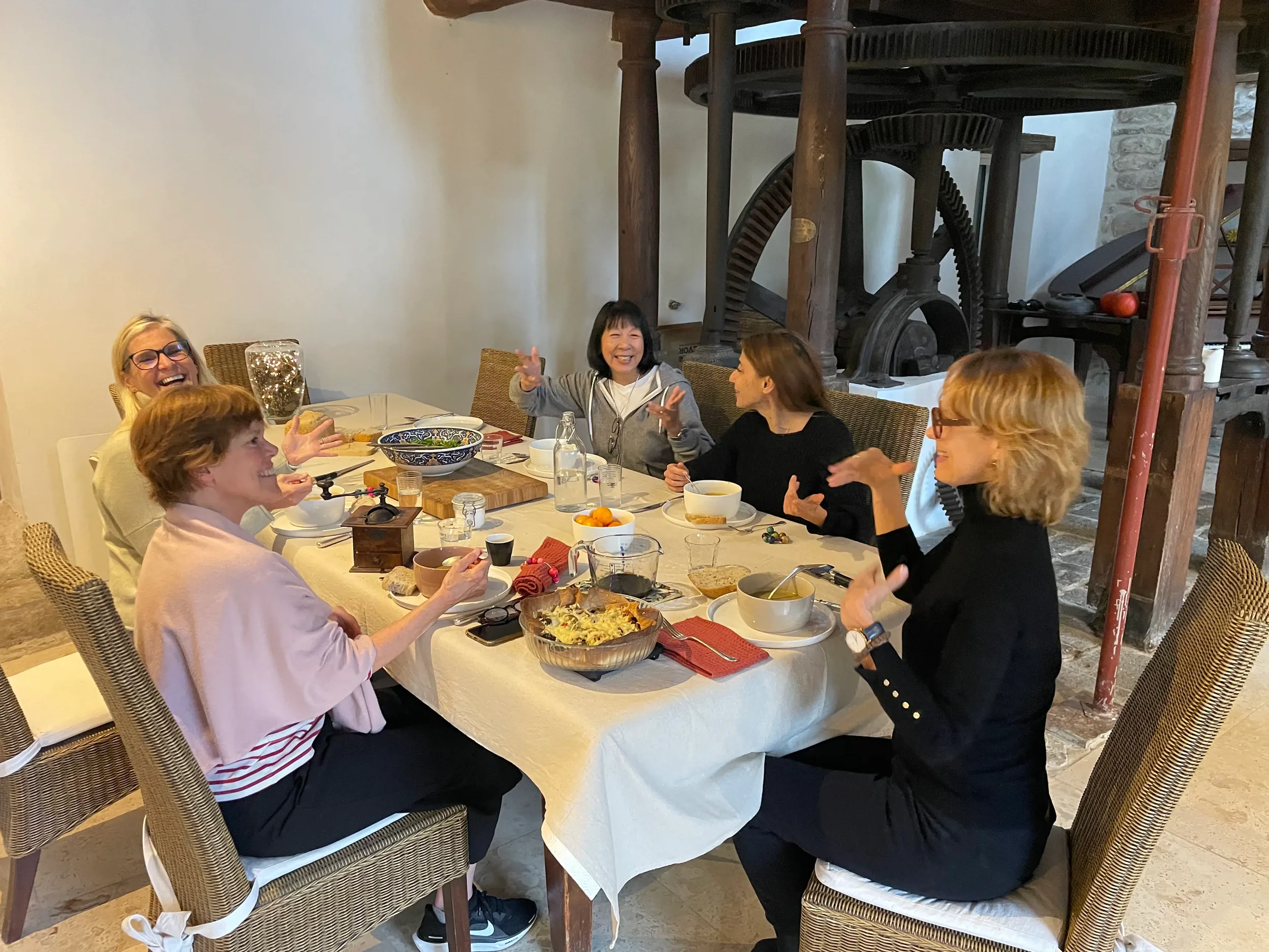 Five women are sitting around a dining table, enjoying a meal and engaged in conversation. The table has various dishes, including a bowl of pasta, a bowl of soup, and some fruit. The background shows vintage machinery and a brick wall.