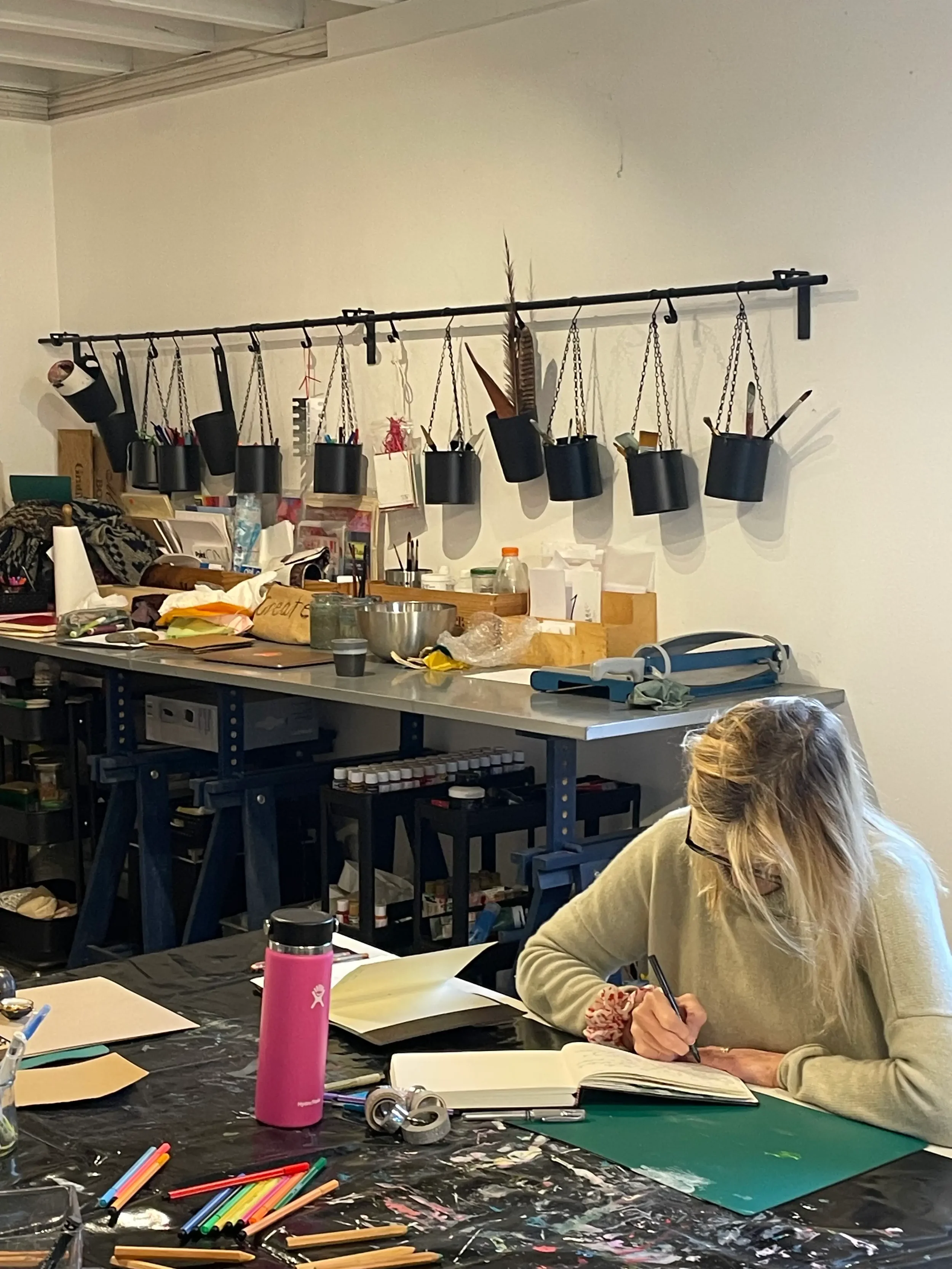 A woman with blonde hair and glasses is sitting at a cluttered desk, writing in a notebook. The workspace has art supplies, a pink tumbler, and various papers. On the wall behind her, there are multiple black hanging containers holding art tools and 