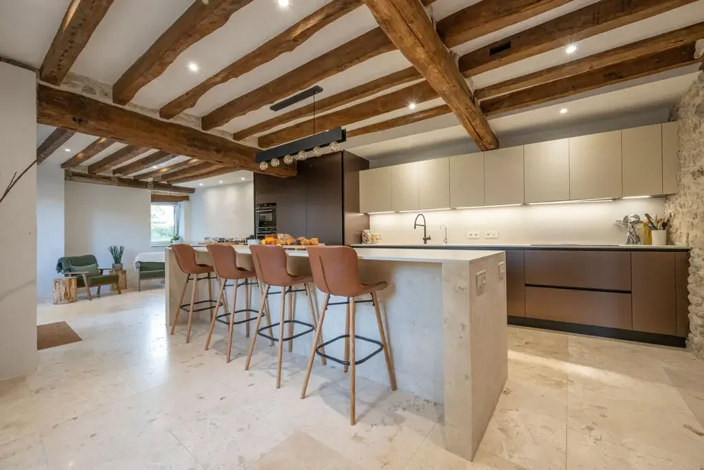 Modern kitchen with a central island with four pink barstools, beige and dark cabinetry, exposed wooden ceiling beams, marble flooring, and a stone wall section.