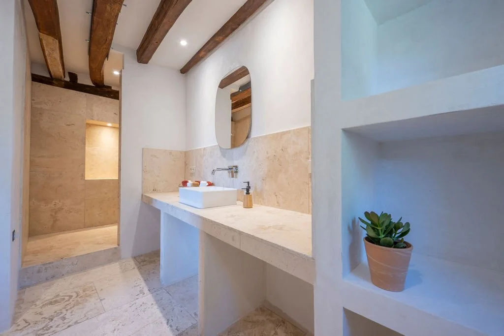 Modern bathroom with white walls, beige stone countertops, and wooden ceiling beams. There is a round mirror above the sink, and a small potted plant on a built-in shelf.