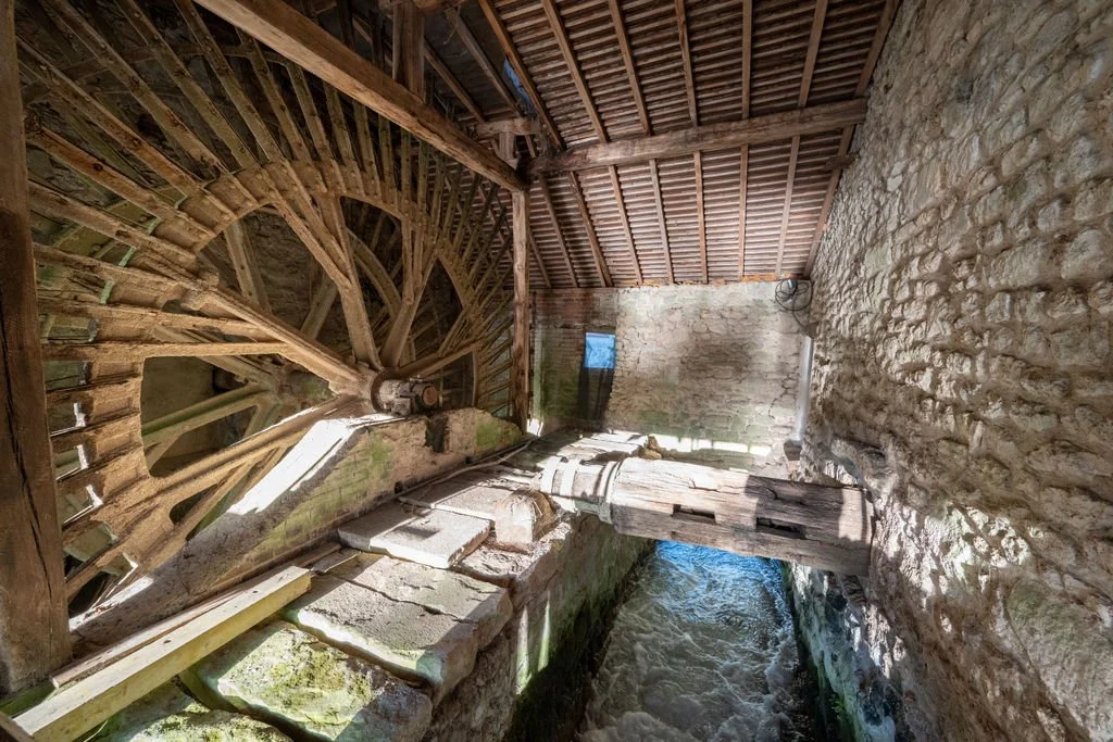 View inside an old stone mill with a wooden water wheel on the left and a flowing stream at the bottom.