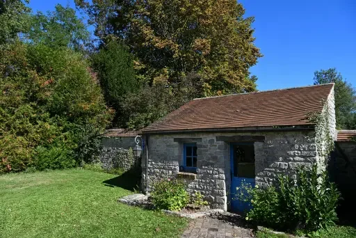 A stone building with a brown roof, blue door, and window, surrounded by green grass and trees.