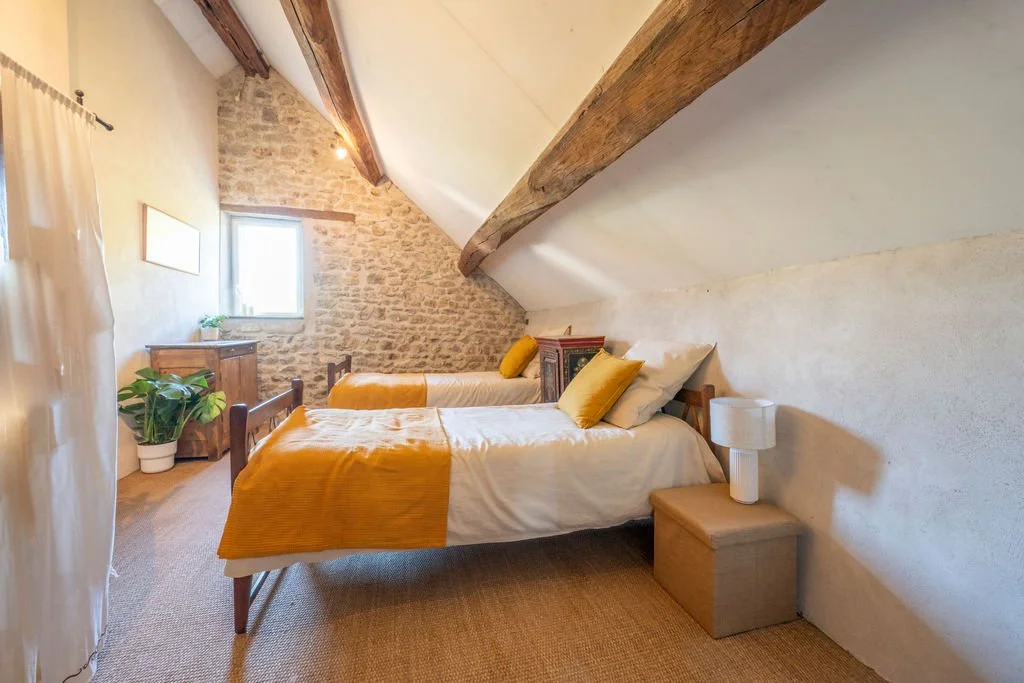 A cozy attic bedroom with two single beds with white and yellow bedding, a small wooden side table with a white lamp, a framed wall art, a potted plant in the corner, a small window, and exposed wooden beams