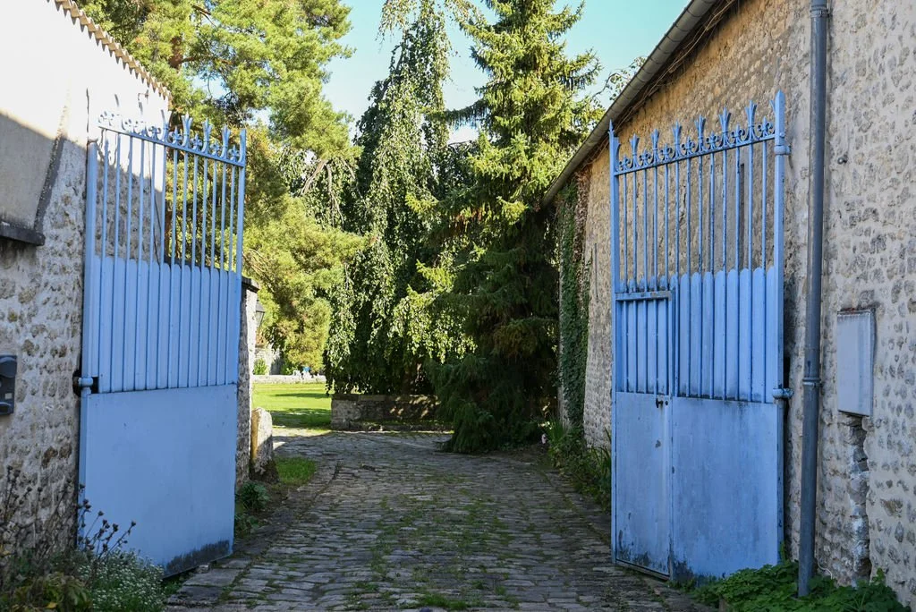 Open blue gate leading to a cobblestone pathway with trees and greenery in the background.