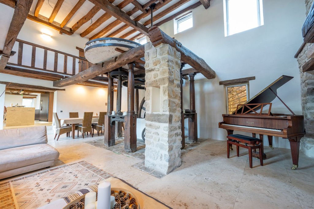 Living room with stone and wood accents, including a grand piano next to a small window, a dining area with a wooden table and wicker chairs, a beige sofa, and decorative candles on a table. Exposed wooden beams are visible on the ceiling, and a mezz