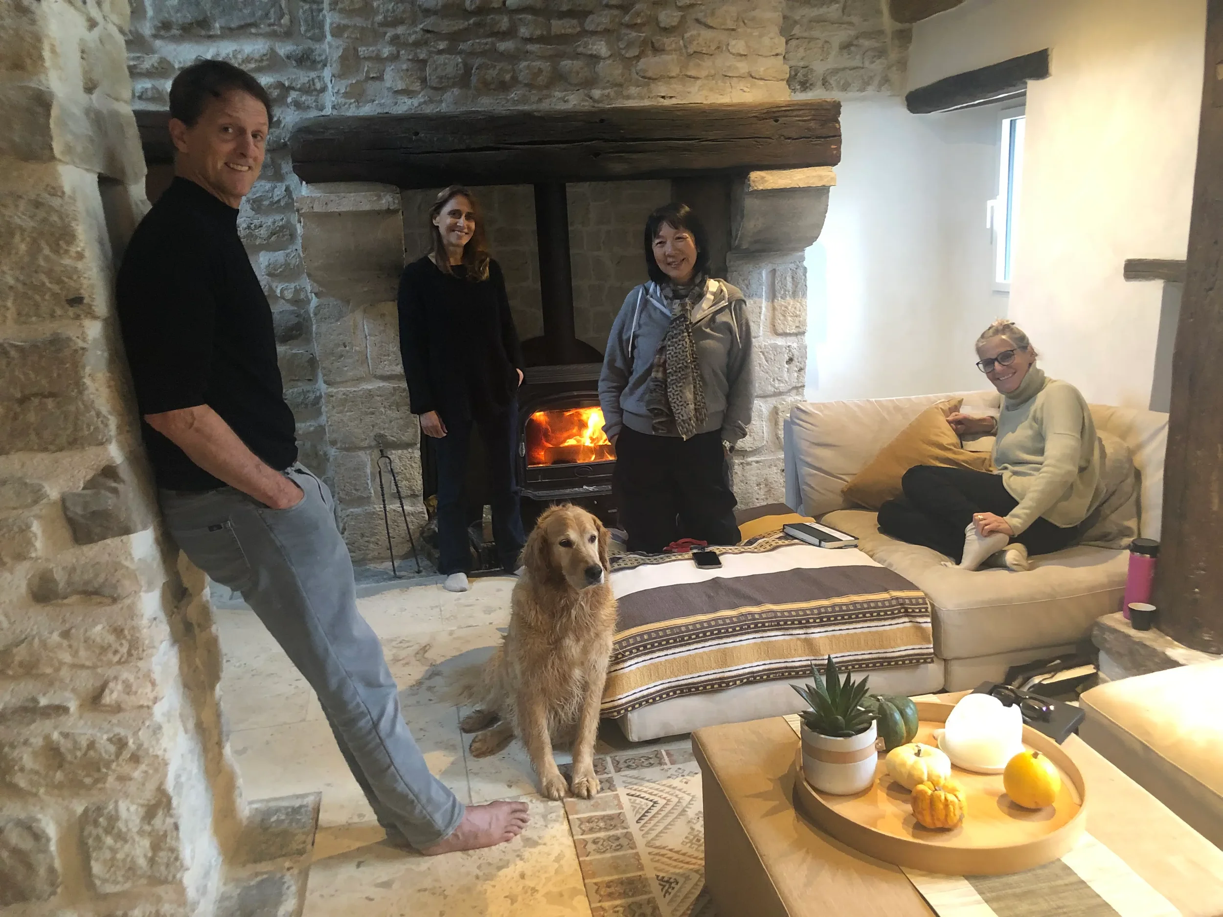 Four women and one man in a living room with a stone fireplace, a dog sitting on the floor, and a sofa with a woman sitting on it. The fireplace has a fire burning, and there are decorative pumpkins and plants on a table in the foreground.
