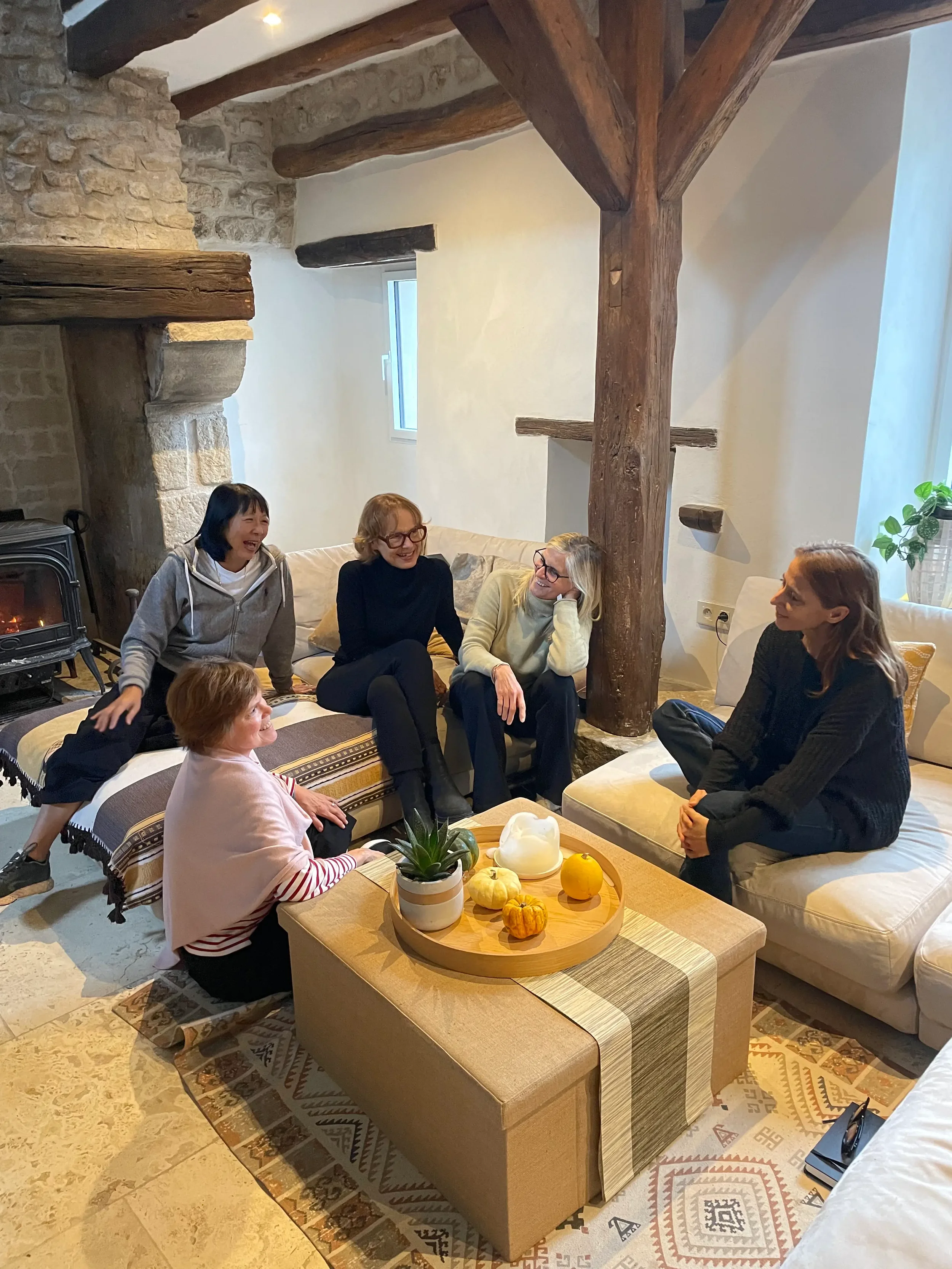 Five women sitting and talking in a cozy living room with wooden beams, a stone fireplace, and decorated with pumpkins and a plant.