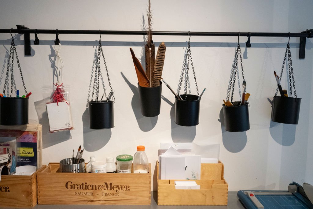 Wall with seven hanging black buckets on chains, each holding art supplies like pens, brushes, and paper. A wooden box below holds bottles and containers, and a central black cup contains feathers and a stick. A paper cutter is partially visible on t