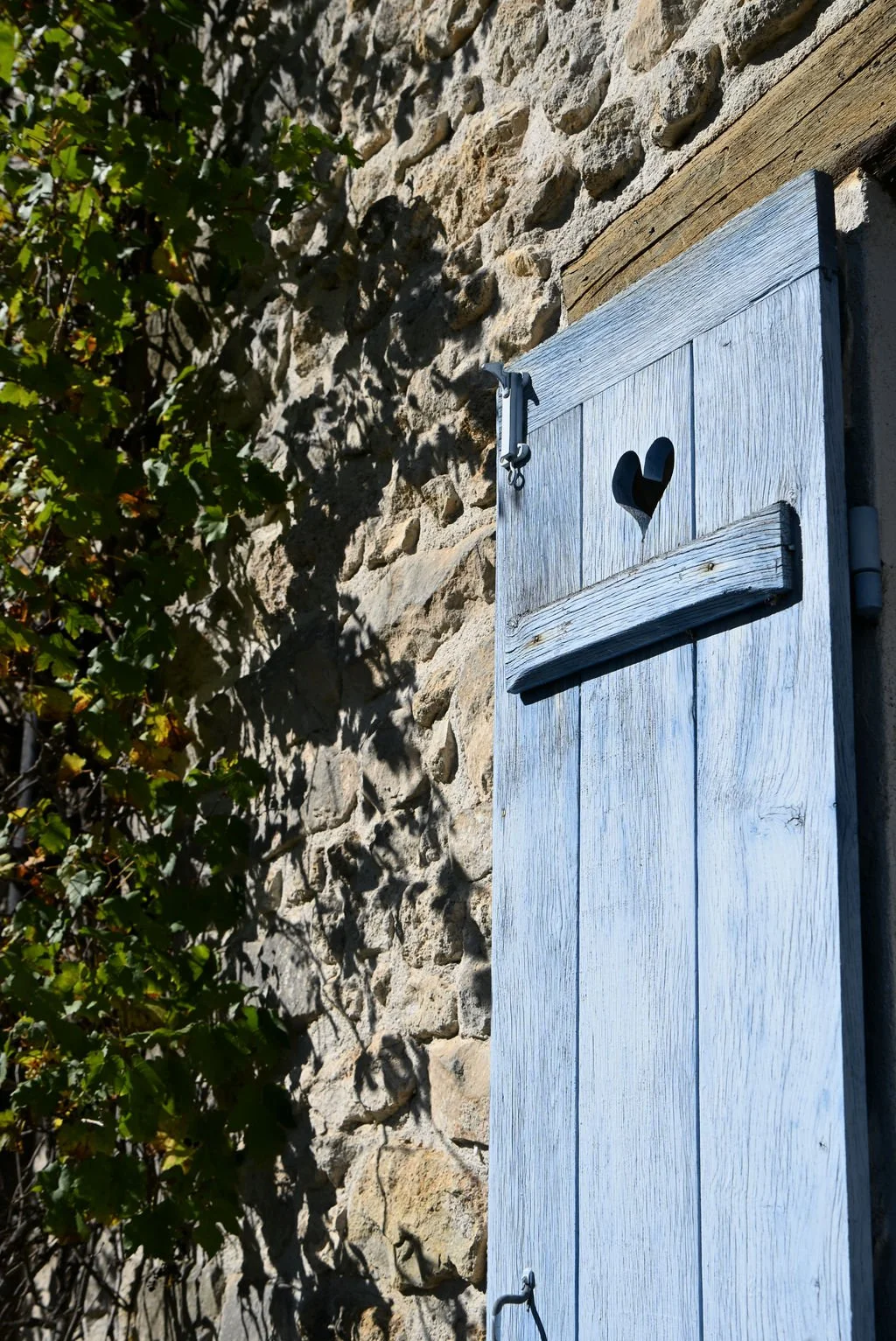 A rustic light blue wooden shutter with a heart-shaped cutout in the center, mounted on a stone wall. Green foliage is partially visible on the left side.