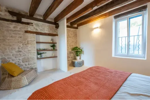 Bedroom with stone wall, wooden ceiling beams, a window with metal bars, a bed with an orange blanket, a yellow pillow, a woven chair, open shelves with plants, and a potted plant on a stand.