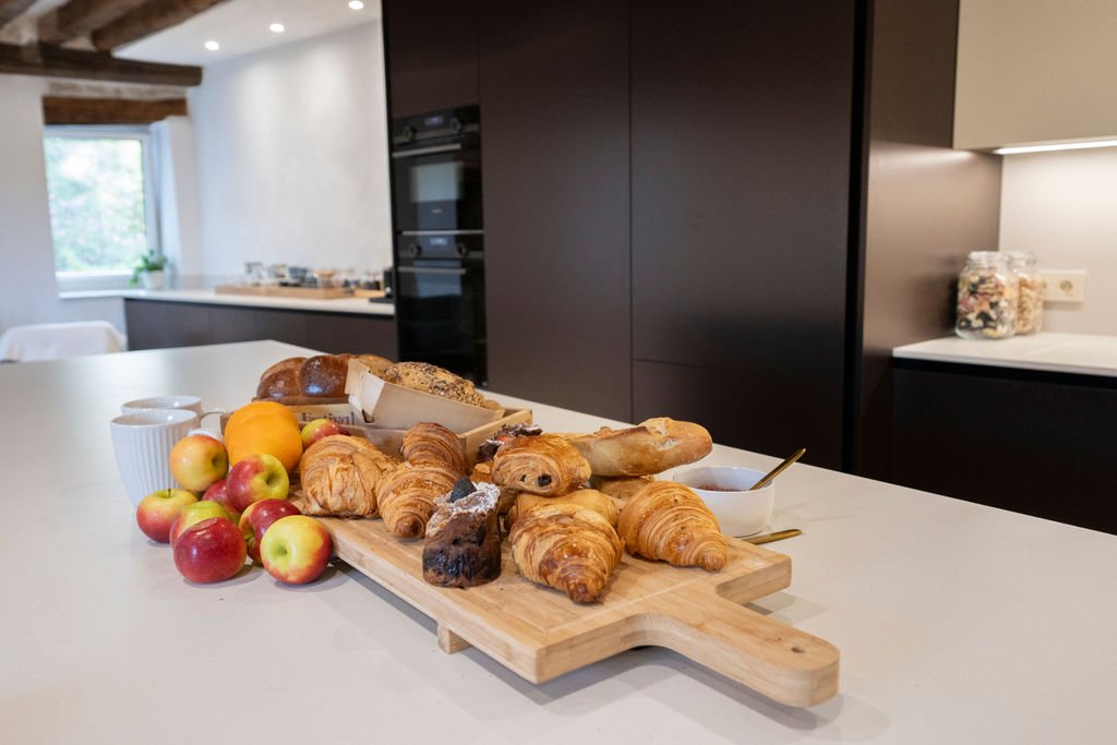 Fresh apples, croissants, bread, and breakfast pastries on a wooden board in a modern kitchen.