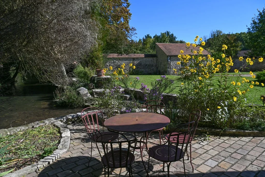 A patio area with a round metal table and four chairs surrounded by lush greenery, yellow flowers, and a small pond on the left side, with a stone building and trees in the background under a clear blue sky.